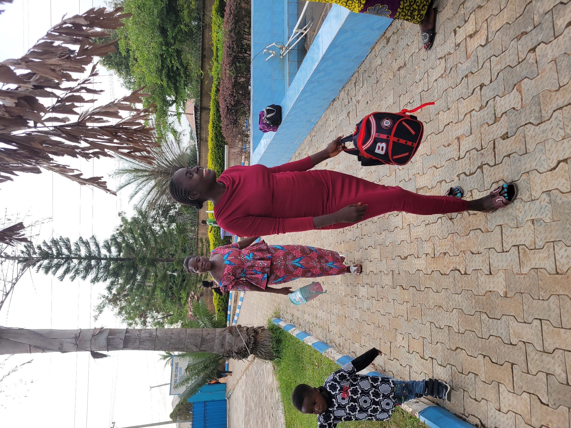 In Jos, Nigeria, the image shows a paved area with interlocking concrete pavers. A woman in a red dress and sandals stands, holding a red and black backpack. Another woman in a patterned dress stands next to her, holding a small bag. A child in a patterned shirt and jeans leans against a blue-and-white painted curb. Behind them is a low wall painted blue, a landscaped area with shrubs, and trees. A second backpack is placed on the wall.