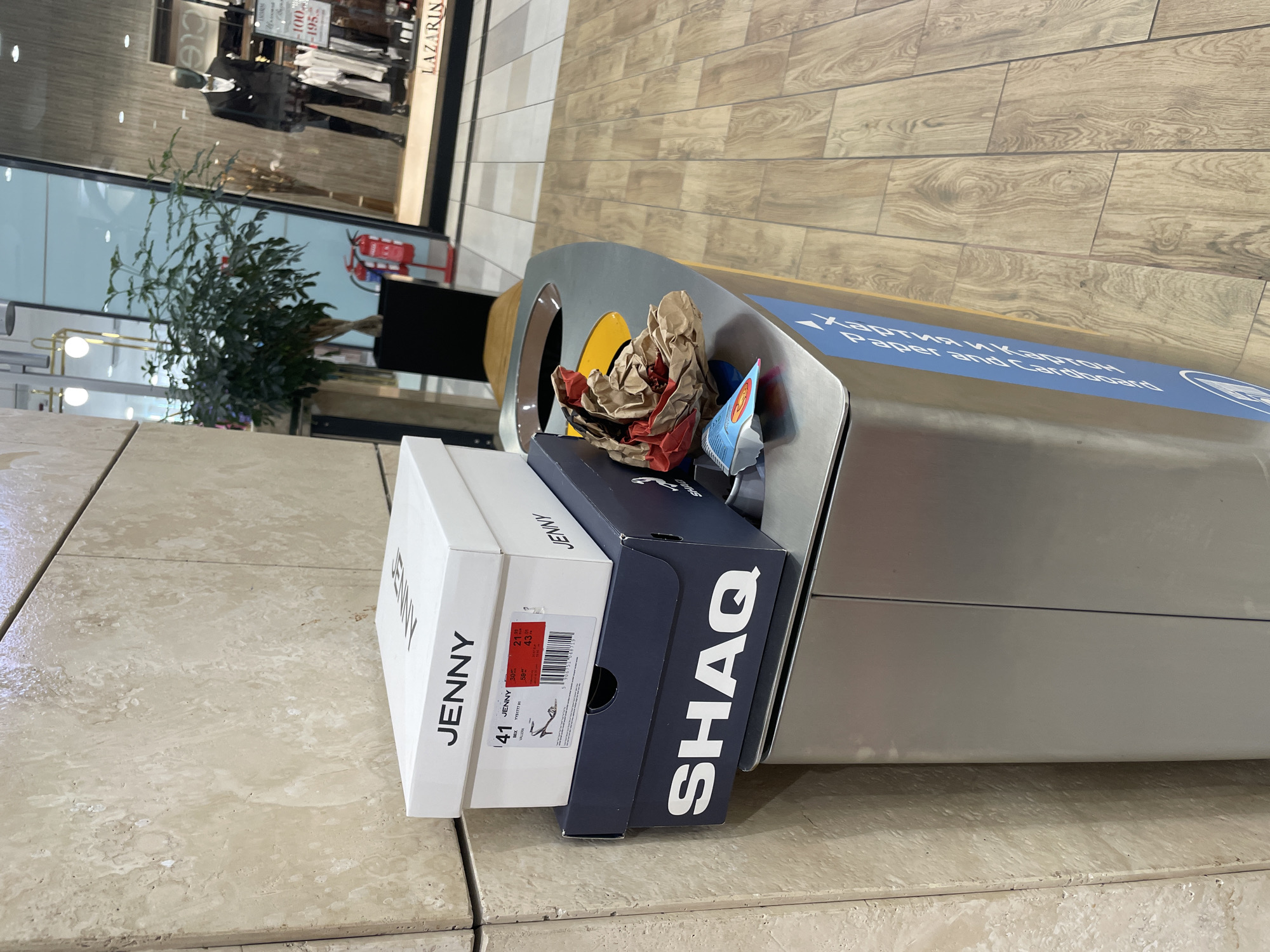 The image captures an indoor commercial environment, identified as a shopping mall in Sofia, Bulgaria, based on visible Bulgarian text and currency.
In the foreground, a stainless steel recycling bin with a rounded top is positioned. The bin has a yellow-rimmed opening and a partially visible blue label on its side indicating "Хартия и картон" (Paper and Cardboard) in white text. Stacked on top of the bin are two shoeboxes. The upper box is white with "JENNY" printed in black, displaying a size 41 and a price label featuring "BGN" currency. Below it is a dark blue shoebox with "SHAQ" printed in large white capital letters. Adjacent to the shoeboxes, on the bin's surface, are crumpled brown paper and a small blue plastic item with a red circular logo.
To the left, a vertical section of a light beige, textured stone-tiled wall is visible. The floor throughout the scene is covered with light-toned, rectangular tiles designed to resemble wood planks, laid in a staggered pattern.
In the mid-ground, a store display window is visible, with the brand name "LAZARINI" written in white at the bottom. Inside, a male mannequin dressed in a black suit, white shirt, and gold bow tie stands, surrounded by clothing racks and sale signs displaying "100,-" and "195,-". A leafy green potted plant in a black rectangular planter is situated between the tiled wall and the store window. Behind the plant and to its right, a red fire extinguisher on a stand is visible near the store's entrance. Reflections of overhead lighting are present in the store window. No individuals are directly visible in the immediate scene.