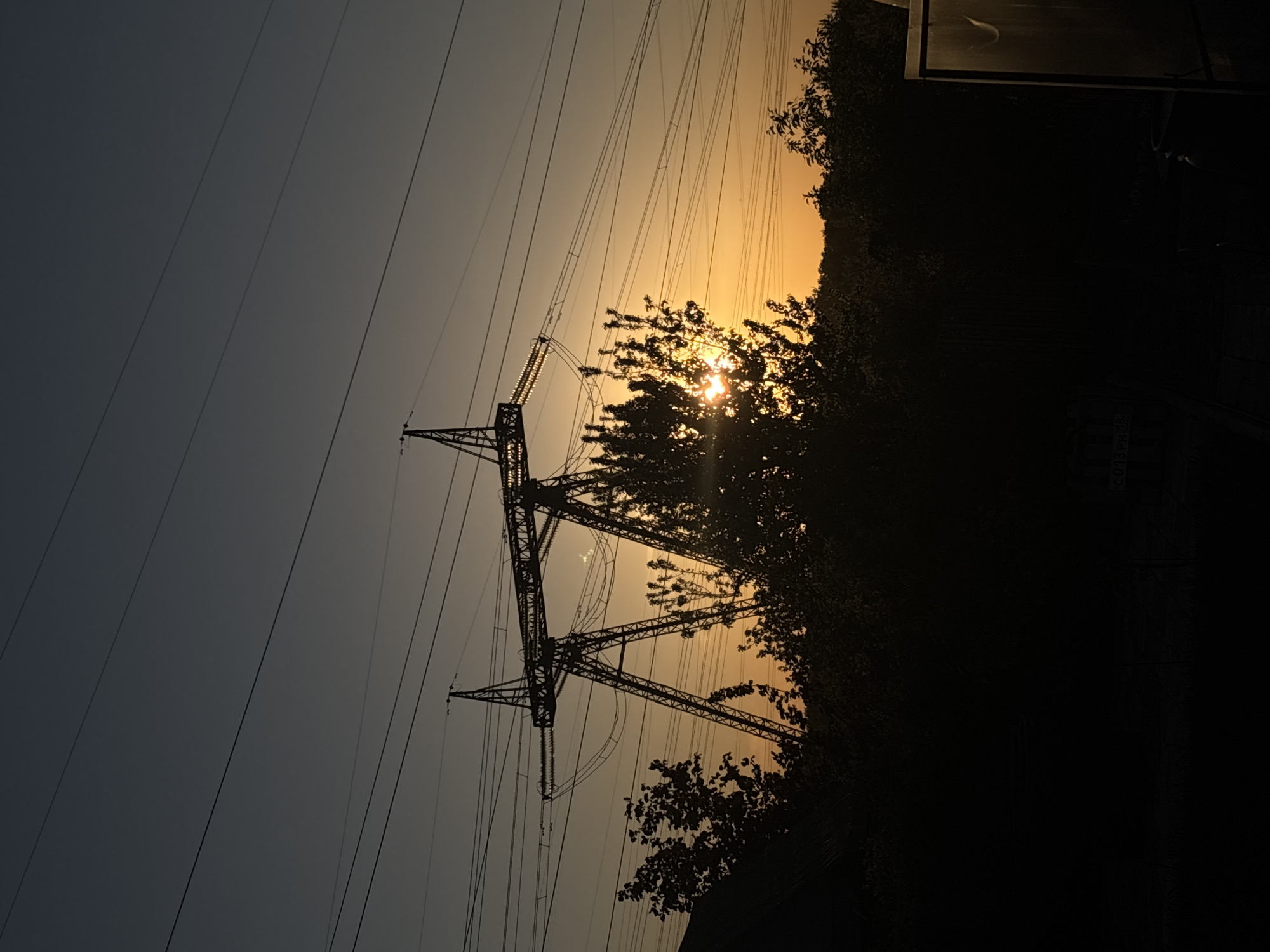 The image shows a high-voltage power transmission tower silhouetted against a setting or rising sun, likely taken in an area with significant vegetation. The power lines extend from the tower towards the left. The right side of the image is dominated by dark trees and a building. The time of day appears to be either early morning or late evening, given the low sun angle and the silhouetted appearance of the objects.
There is no information in the image to support the suggestion it was taken in the city of Chaikovsky, Russia.