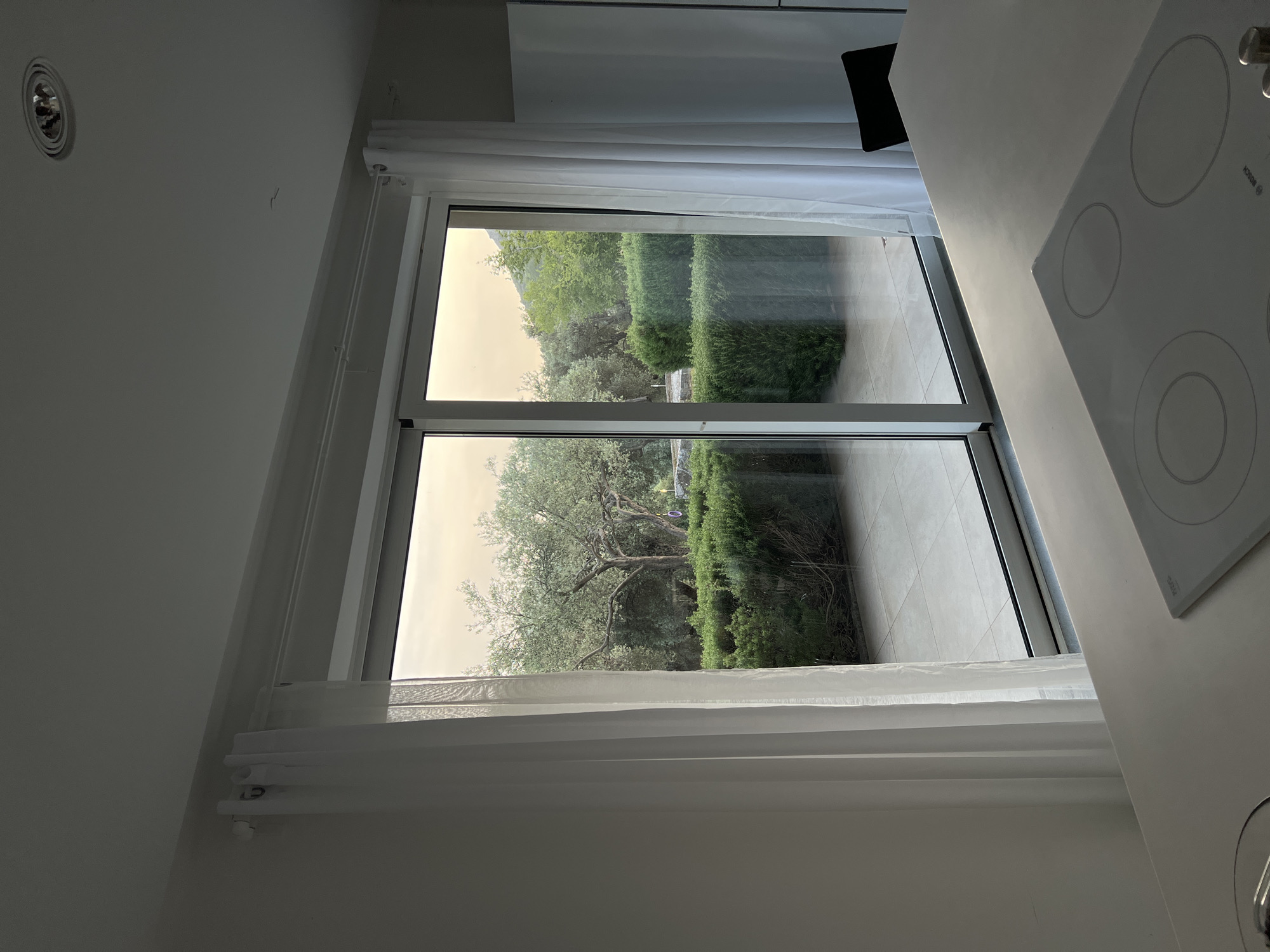 A vertically-oriented medium shot depicts a view through a closed sliding glass door in what appears to be an interior space. The sliding door is framed by white curtains on either side. Outside, a manicured garden is visible, featuring a large tree and neatly trimmed hedges. The lighting outside suggests an overcast or hazy day.
The interior space is minimalistic, with white walls and a white countertop visible on the right side of the frame. A white electric stovetop with four burner circles is partially visible on the countertop. The scene implies a clean and modern aesthetic. The location may be near Ivanovići, Montenegro.