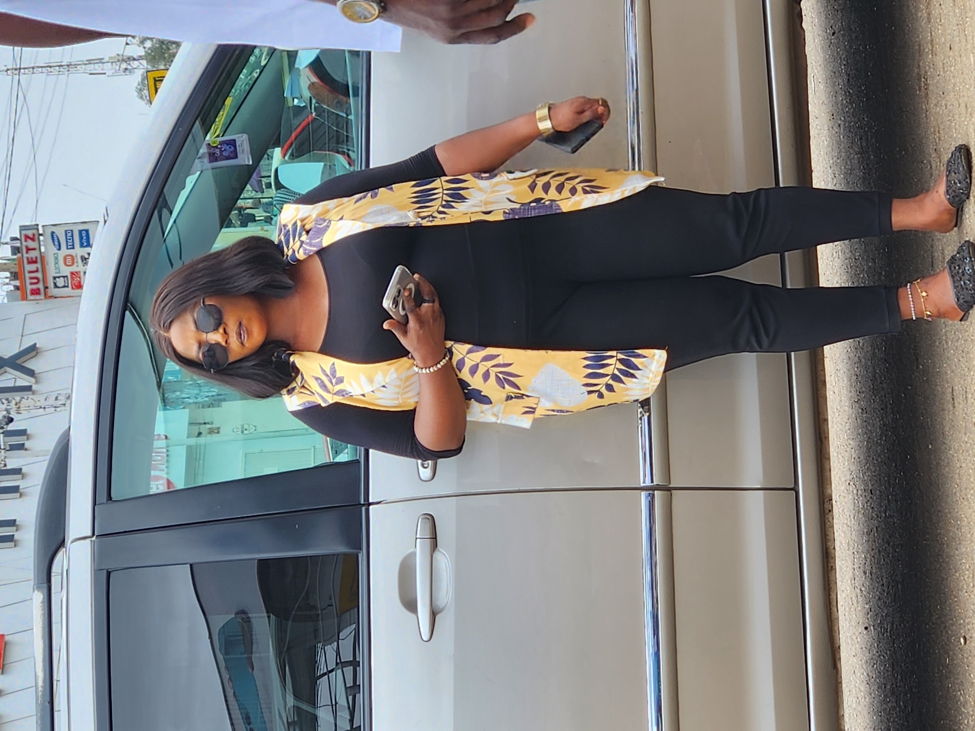 A woman stands next to a silver van. She wears black sunglasses, a black shirt and pants, and a yellow and white floral print vest. She holds a phone in each hand. Another person stands to her right with their arm visible. A shop sign is visible in the background, with the brand names "BULETZ," "LG/ORIX," "SAMSUNG," "TECHO," and "VIVO." The location is in the city of Unknown, Unknown.