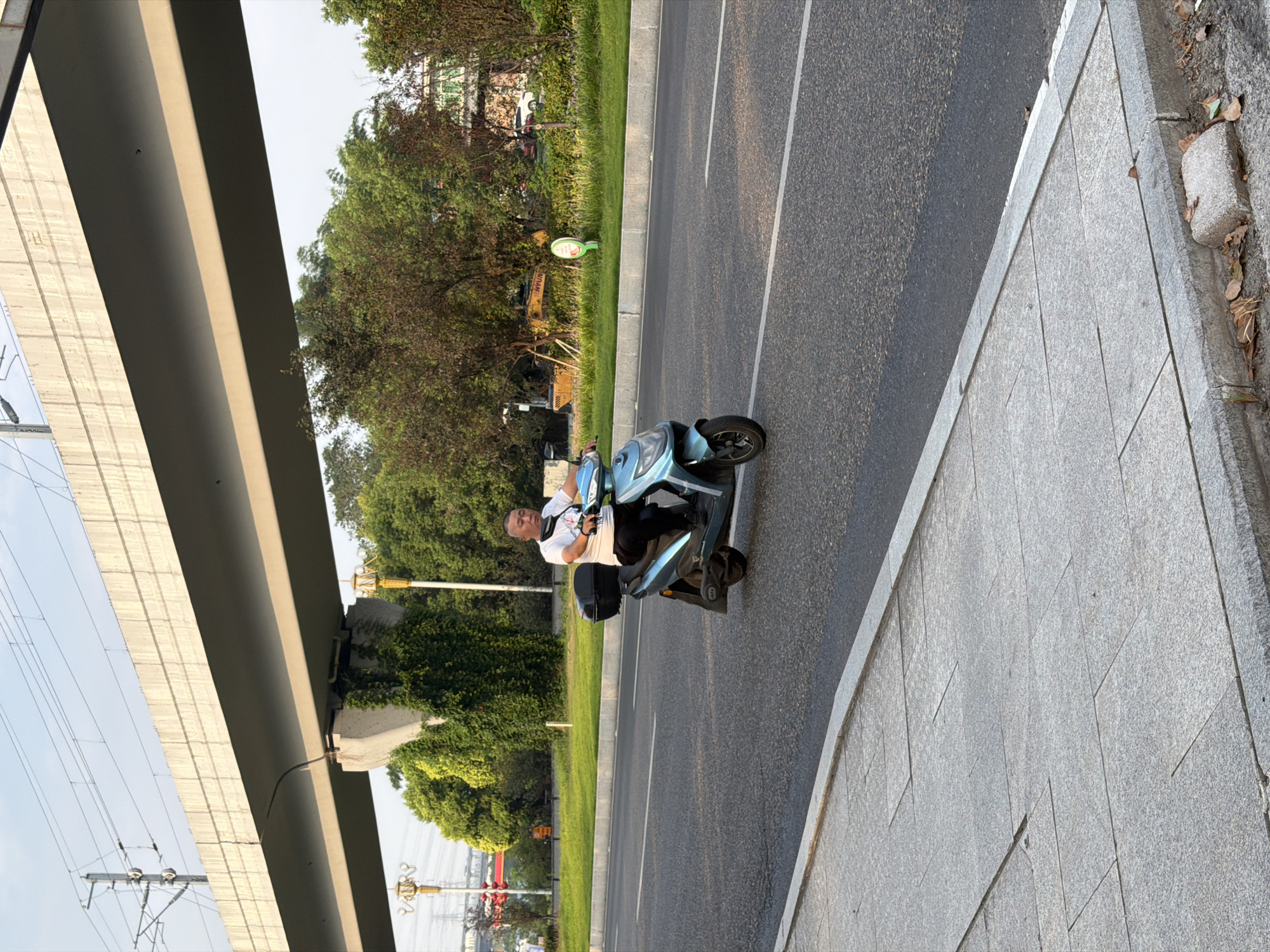 In Jiangjin, China, a man is riding a light blue electric scooter on a paved road. He is wearing a white t-shirt and dark pants, with a black helmet secured around his neck. The scooter has a black storage box on the back. The road is flanked by a concrete sidewalk on the left and a grassy median with trees and vegetation on the right. An elevated concrete structure, possibly a highway or railway bridge, stretches across the upper portion of the image. The sky is clear and blue.