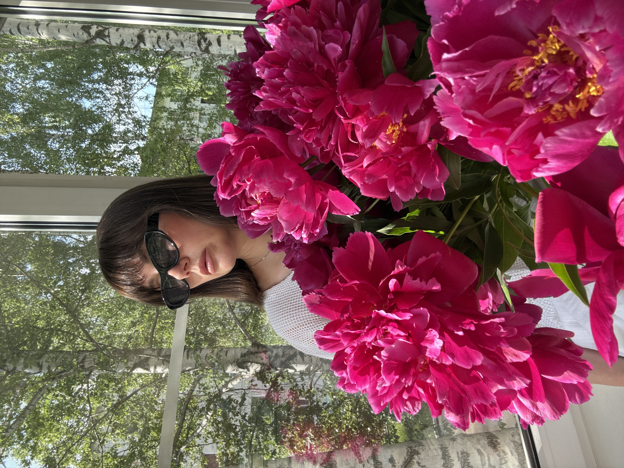 A medium shot captures a woman with dark hair and bangs, wearing black cat-eye sunglasses, standing in front of a window with a view of birch trees and a building. She is holding a large bouquet of bright pink peonies, which fill the foreground of the image. The woman is wearing a white, loosely knit sweater. The scene appears to be an indoor setting near a window, possibly in the city of Chaikovsky, Russia, given the birch trees visible outside. The lighting suggests it is daytime, with sunlight filtering through the window and illuminating the flowers.