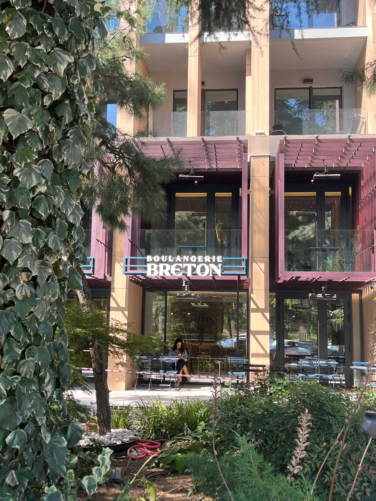 The image shows a bakery called "BOULANGERIE BRETON" located in Tbilisi, Georgia. The bakery has a modern facade with large glass windows and doors, and an outdoor seating area. The exterior is decorated with wooden panels and maroon colored architectural elements. In front of the bakery, there are several outdoor tables and chairs. A woman is sitting at one of the tables, looking at her phone. The foreground of the image is filled with lush green foliage, including a large ivy-covered tree and other plants. The lighting suggests it is a sunny day.