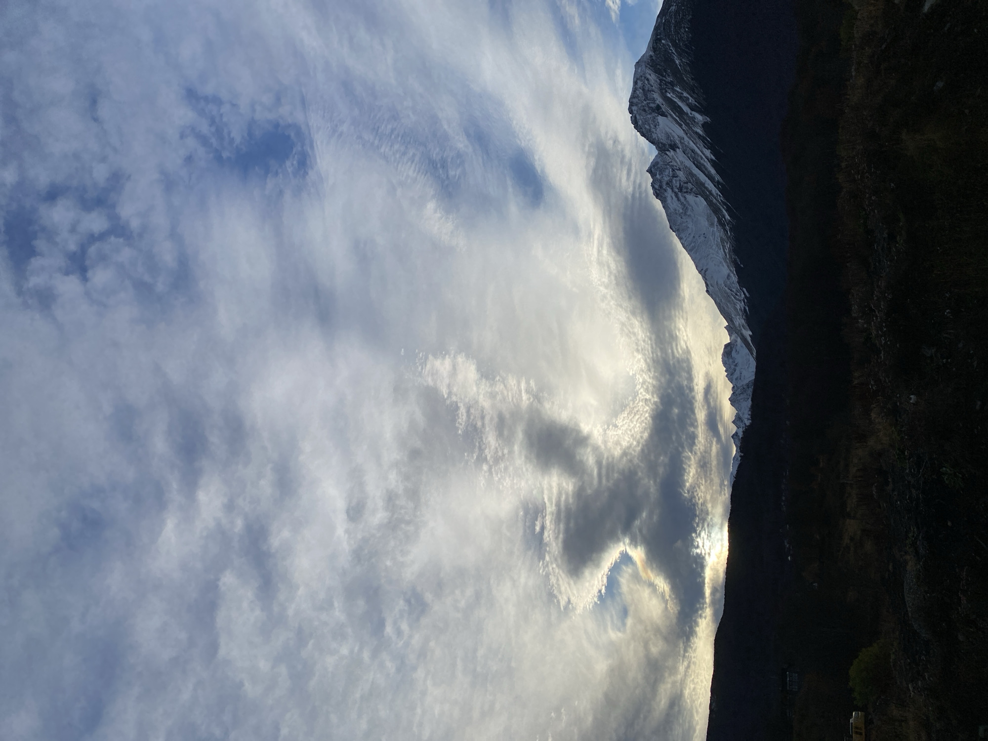 The image shows a mountain range with snowy peaks in Ushuaia, Argentina, with a sky filled with white and gray clouds. A small section of land to the right of the mountain range is covered in dark vegetation. The image seems to be taken during the day.