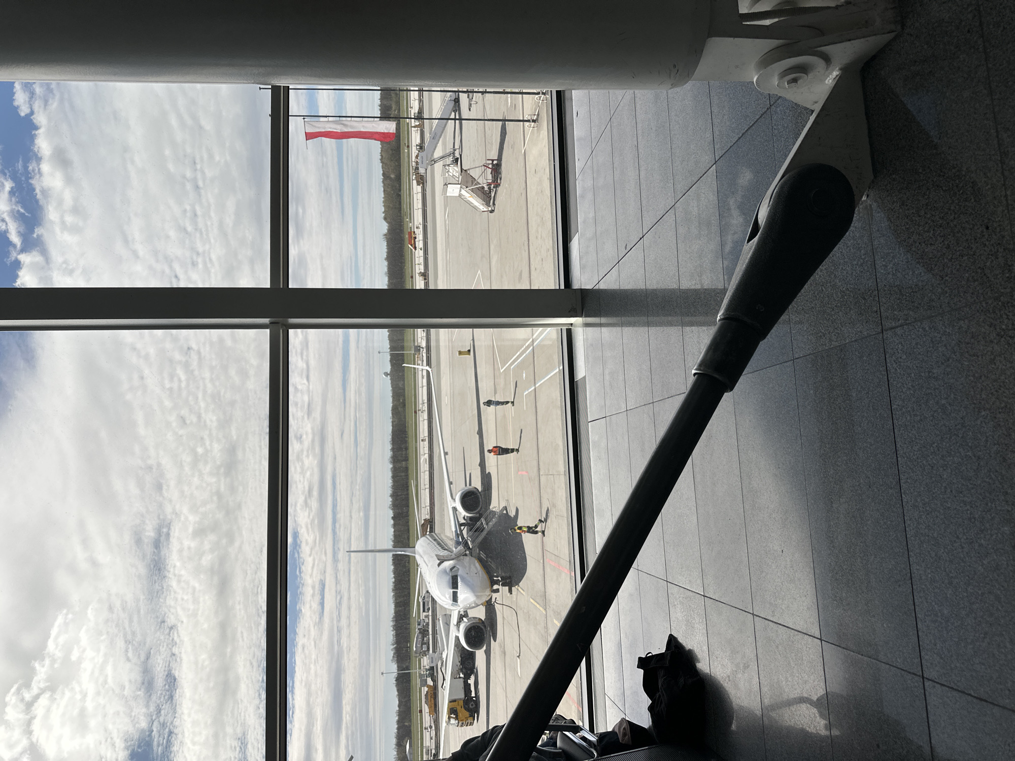 A view from inside the Wrocław Airport terminal in Wrocław, Poland, shows an airplane parked on the tarmac, viewed through a window. Airport workers are visible on the ground near the airplane. A Polish flag is visible in the background. A structural support beam is in the foreground inside the terminal. A bag is on the floor near the base of the support beam. The sky is partly cloudy.