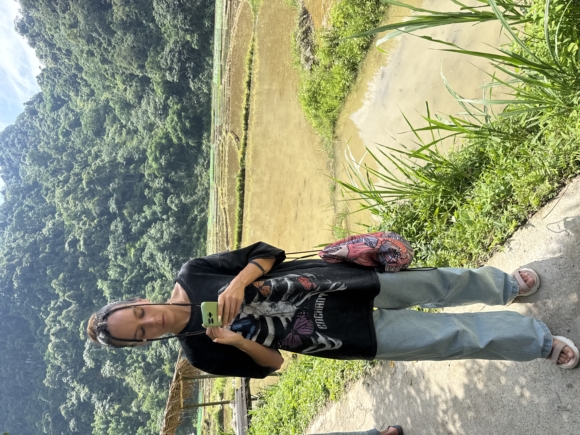 A woman stands on a concrete path with a phone in her hands, looking down at it. She is wearing a black t-shirt with a skeleton and butterfly graphic, light blue baggy jeans, and pink sandals. A patterned bag is slung over her right shoulder. Behind her, terraced rice fields filled with water extend towards a lush, green, tree-covered mountain under a bright sky. A small thatched-roof structure is visible to the left of the woman. The scene is set in Pù Luông Commune, Vietnam.