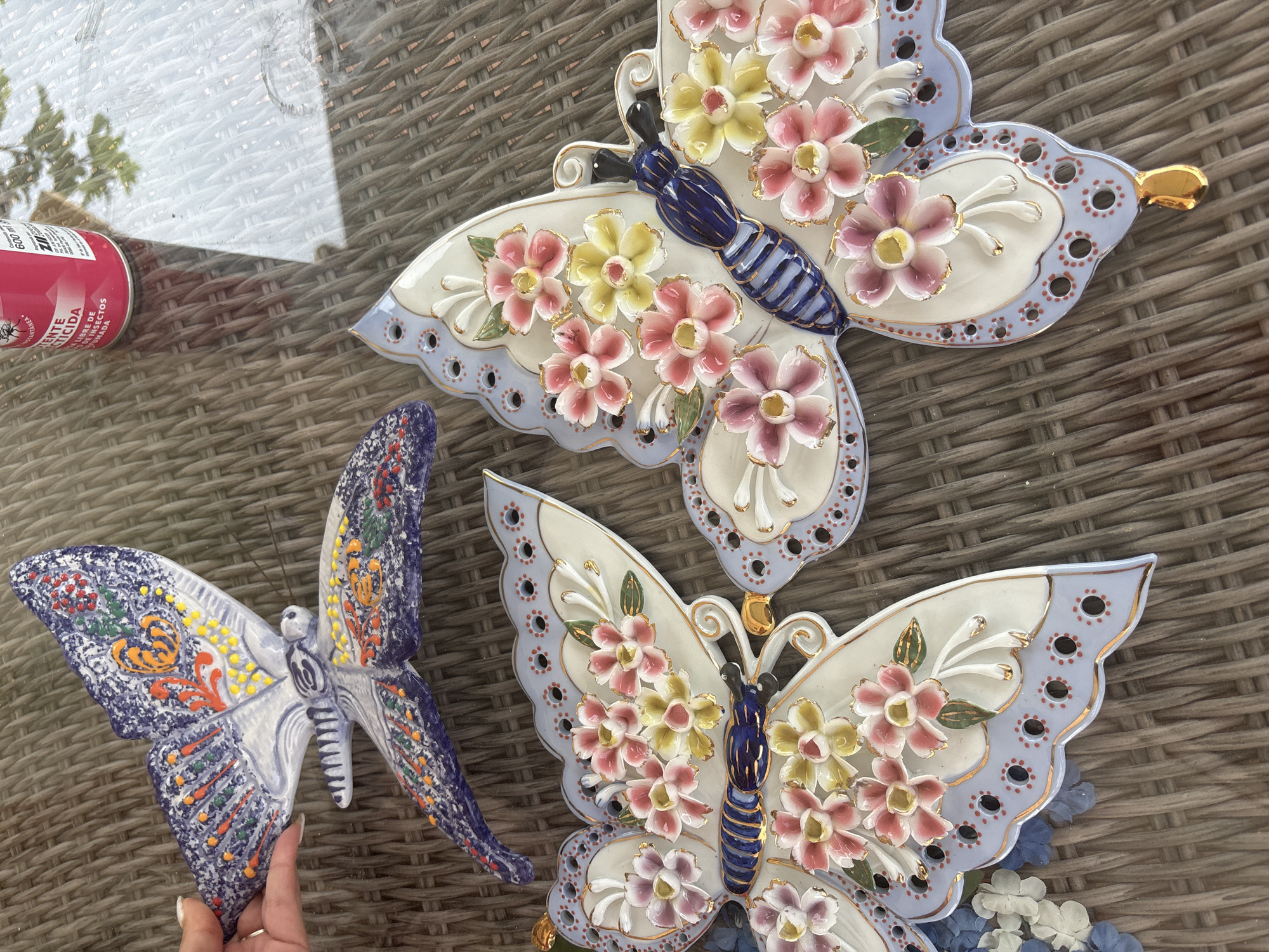 Three ceramic butterfly decorations are arranged on a woven outdoor table surface. Two of the butterflies, larger and more ornate, feature intricate floral designs in pink, yellow, and white, with blue and gold accents. The third butterfly, positioned to the left and partially held by a hand with a silver ring, has a more abstract, painted design in shades of blue, purple, orange, and red. A pink spray can with Spanish text is visible in the upper left corner. The background is a blurred reflection on a glass surface, suggesting an outdoor setting. The overall scene is a close-up, top-down view of decorative items.
