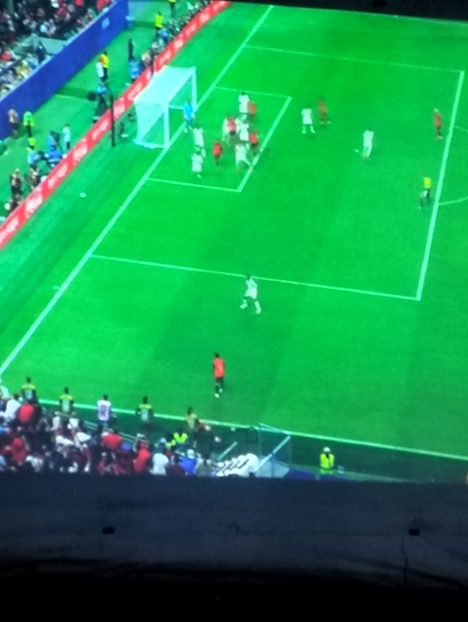 The image depicts a soccer match in progress. The field is green, with white lines marking the boundaries and penalty areas. A white goalpost is visible on the left side of the frame. Several players in white and red jerseys are on the field. In the foreground, a crowd of spectators is visible, many wearing red and white. The scene appears to be captured from a high vantage point, likely a stadium. There is no information within the image to suggest the location is Payagaboh, Indonesia.