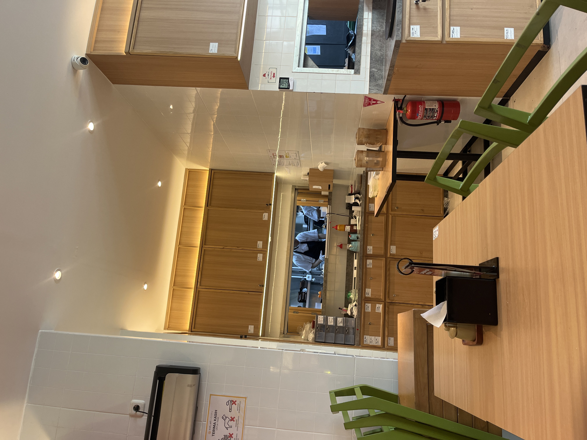 This indoor shot depicts a scene in what appears to be a restaurant or food establishment in Lhokseumawe, Indonesia. The perspective is from a low angle, tilted upward. The walls are covered in white tiles. On the left wall, there's a hand dryer and a sign in Bahasa Indonesia. Above, there are built-in wooden cabinets and recessed lighting.
The focus shifts to the center, where a window reveals a view into the kitchen. Inside, two individuals wearing white tops and dark aprons appear to be working. Various cooking equipment and supplies are visible within the kitchen. Above the kitchen window are more wooden cabinets.
To the right of the image, there's a wooden table with green chairs. A tissue dispenser is mounted on the side of the table. A fire extinguisher is mounted on the wall, also towards the right. The general impression is that of a functional and clean environment.