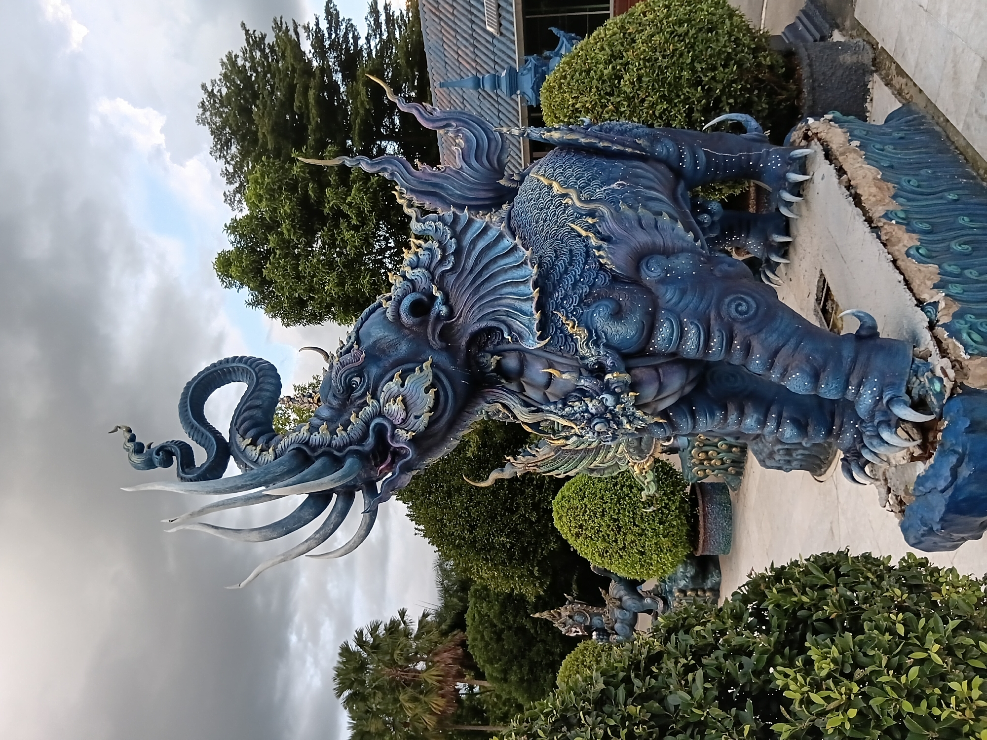 The image depicts a striking, elaborately decorated blue statue of a mythical elephant-like creature with multiple tusks and a serpentine tail, prominently positioned outdoors. The statue is adorned with gold accents and intricate details. It stands on a raised, broken platform with decorative blue wave-like patterns at its base. Surrounding the statue are meticulously manicured green bushes and trees, with a few palm trees visible in the background. In the distance, a building with a gray tiled roof and blue architectural elements can be seen. The sky is overcast with dramatic clouds. The statue and its setting suggest a location within a temple or a cultural attraction in Chiang Rai City Municipality, Thailand, likely the Wat Rong Suea Ten (Blue Temple) given its distinctive blue color and artistic style. No people are visible in the frame. The overall scene is one of impressive artistry and a somewhat somber atmosphere due to the cloudy sky.
