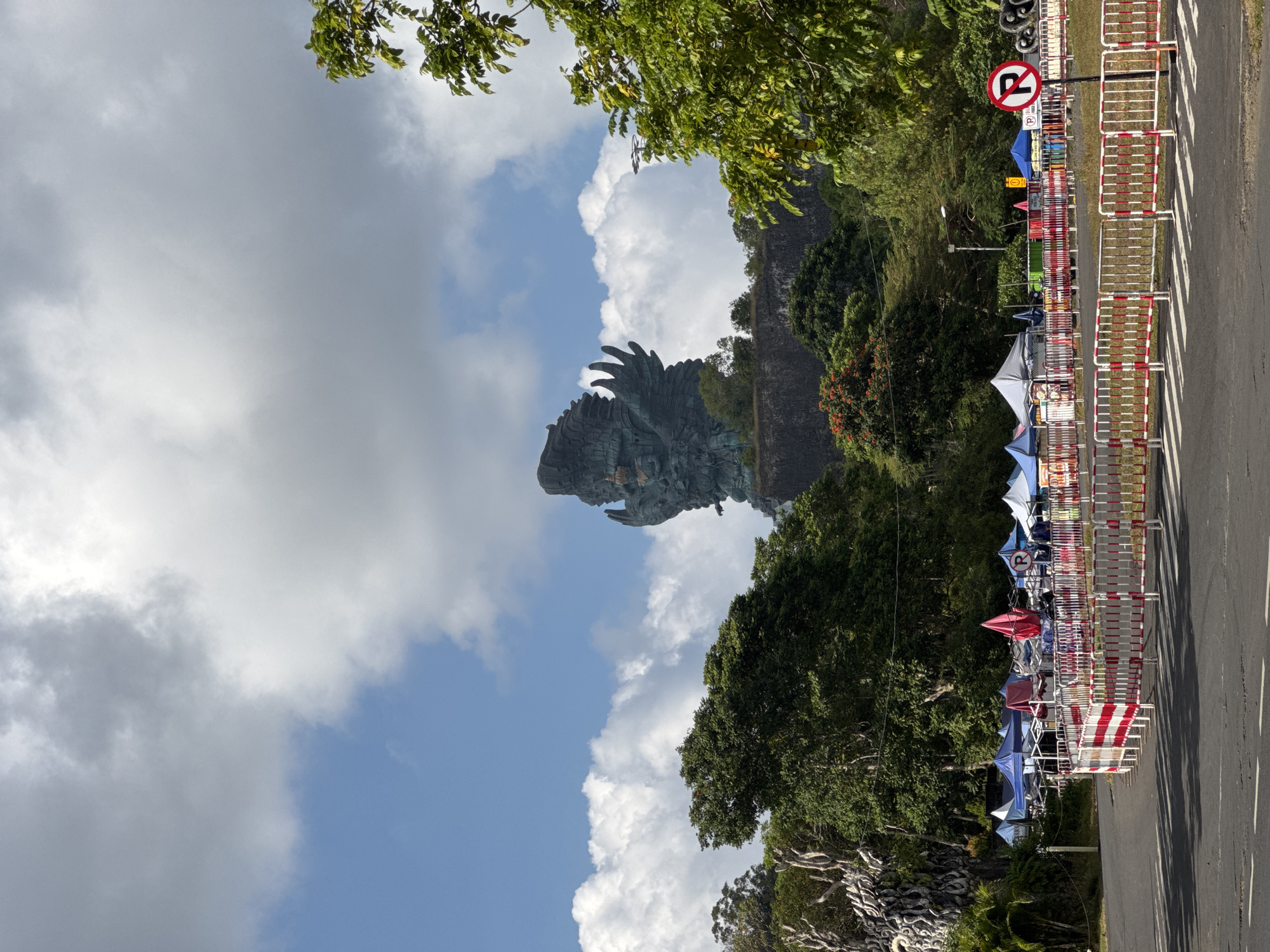 The image displays an outdoor scene, rotated 90 degrees counter-clockwise, capturing a large statue and its surroundings under a partly cloudy sky. The primary subject is a monumental, dark-colored statue of a bird-like figure, resembling the Garuda, positioned towards the upper-left of the frame, towering over a landscape of lush green trees. The statue's intricate details, particularly its wings and head, are visible against a bright blue sky dotted with white and grey clouds.
In the right portion of the image, a paved road stretches along, separated from a pedestrian or market area by a series of red and white striped metal barriers. Beyond these barriers, several market stalls or vendor tents with colorful canopies (including blue, red, and white) are set up, suggesting an active commercial or tourist zone. A distinct circular "no parking" sign is prominently featured near the road and barriers. The sign has a red circle, a white background, and a black letter 'P' with a diagonal line through it, indicating a parking restriction. Below the 'P' symbol, the word "PARKING" is also visible.
The scene appears to be well-lit by natural sunlight, indicating it is daytime, likely midday or early afternoon. The abundance of green foliage and the presence of tourist-oriented stalls suggest a park or cultural attraction. The image conveys a sense of a vibrant public space, likely the Garuda Wisnu Kencana Cultural Park in Kuta Selatan, Indonesia, given the context.