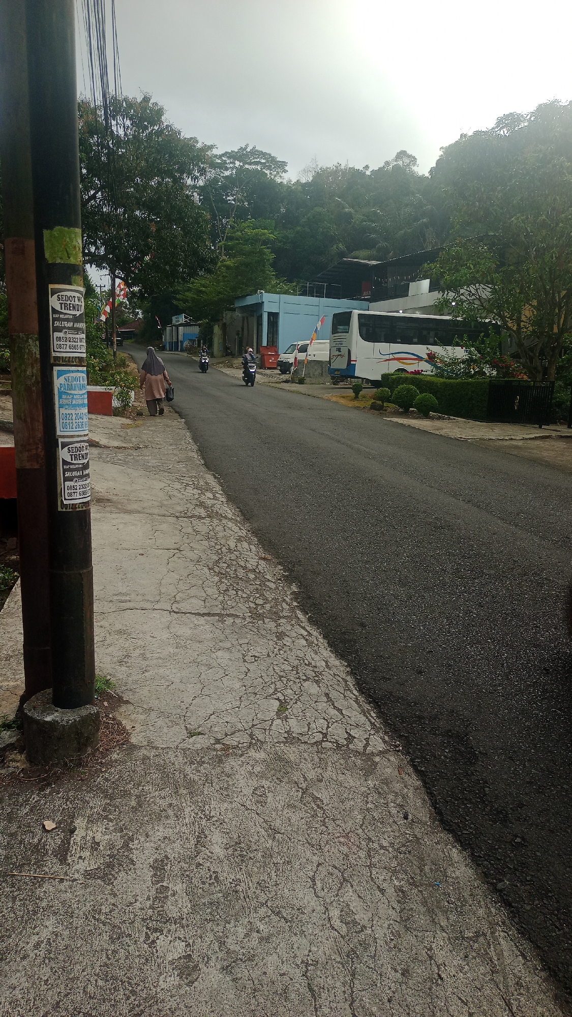 The image depicts a street scene in Karyamandala, Indonesia. On the left side of the road, a utility pole is covered with several stickers, some advertising "SEDOT WC TRENDY" and phone numbers. A cracked concrete sidewalk runs alongside the pole, with a woman in a hijab walking away from the camera on it. Two motorcycles are also visible on the road, one with a rider and another parked.
The road itself is paved and appears to be in moderate condition, with some patches of wear. On the right side of the road, a white bus is parked, with a smaller white van to its left. Behind the vehicles, a light blue building is visible, along with more vegetation and a hillside covered in trees. The sky is overcast and bright, suggesting either early morning or late afternoon. The overall impression is of a quiet, somewhat utilitarian street in a suburban or semi-rural area.