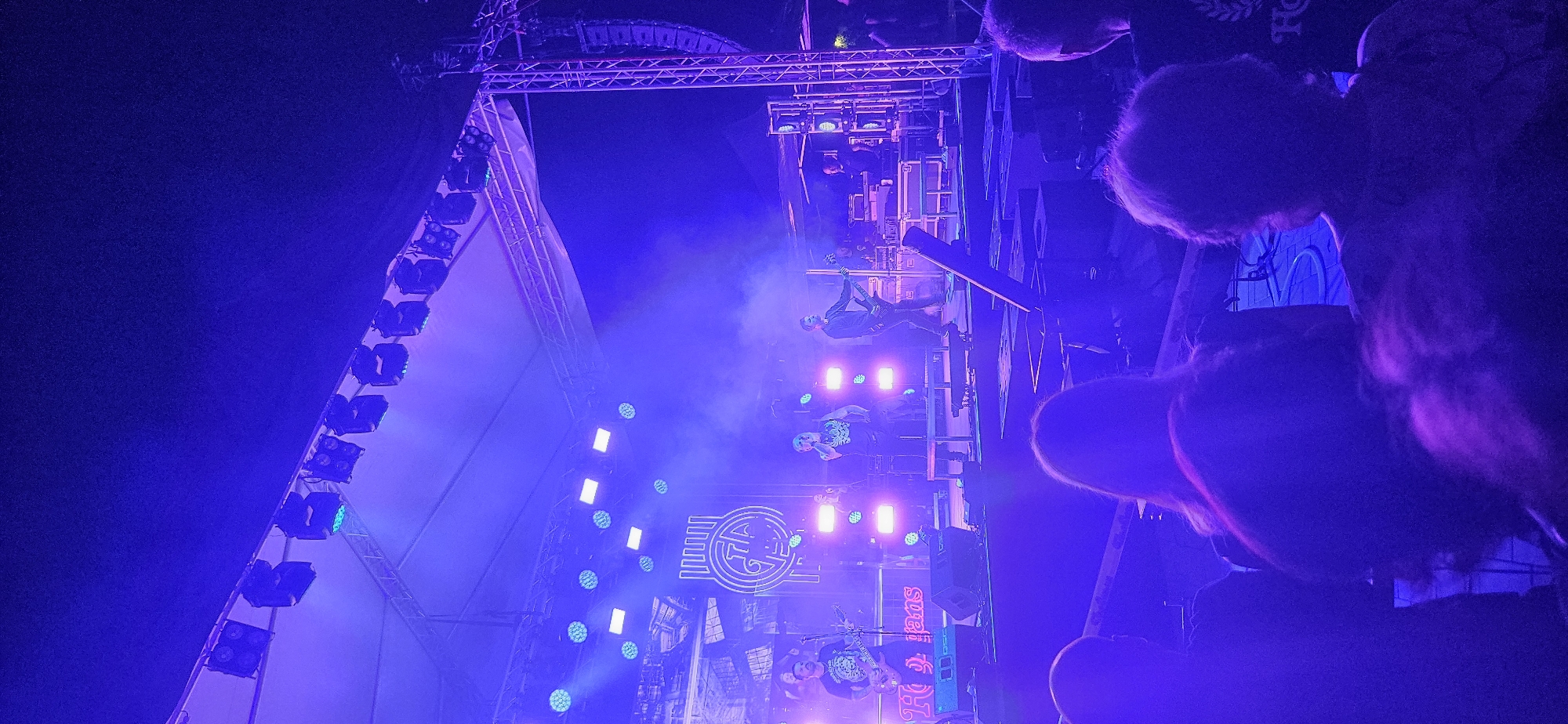 The image depicts an outdoor concert at night, bathed in intense blue and purple stage lighting. A band is performing on a raised stage, with several members visible. One vocalist stands center stage, holding a microphone, while another member plays a guitar to the left. A third person is also visible on stage playing an instrument. The band's logo, a stylized circular emblem with vertical lines, is displayed prominently behind them, along with the text "Hogyan". The stage is equipped with multiple speakers and lighting rigs. In front of the stage, the audience is visible from behind, with many heads and shoulders in the foreground, all illuminated by the stage lights. The overall atmosphere is that of a lively musical performance. While the image does not explicitly state the location, the presence of the text "Hogyan" strongly suggests the concert is taking place in or near Hévíz, Hungary, as "Hogyan" is a Hungarian word.