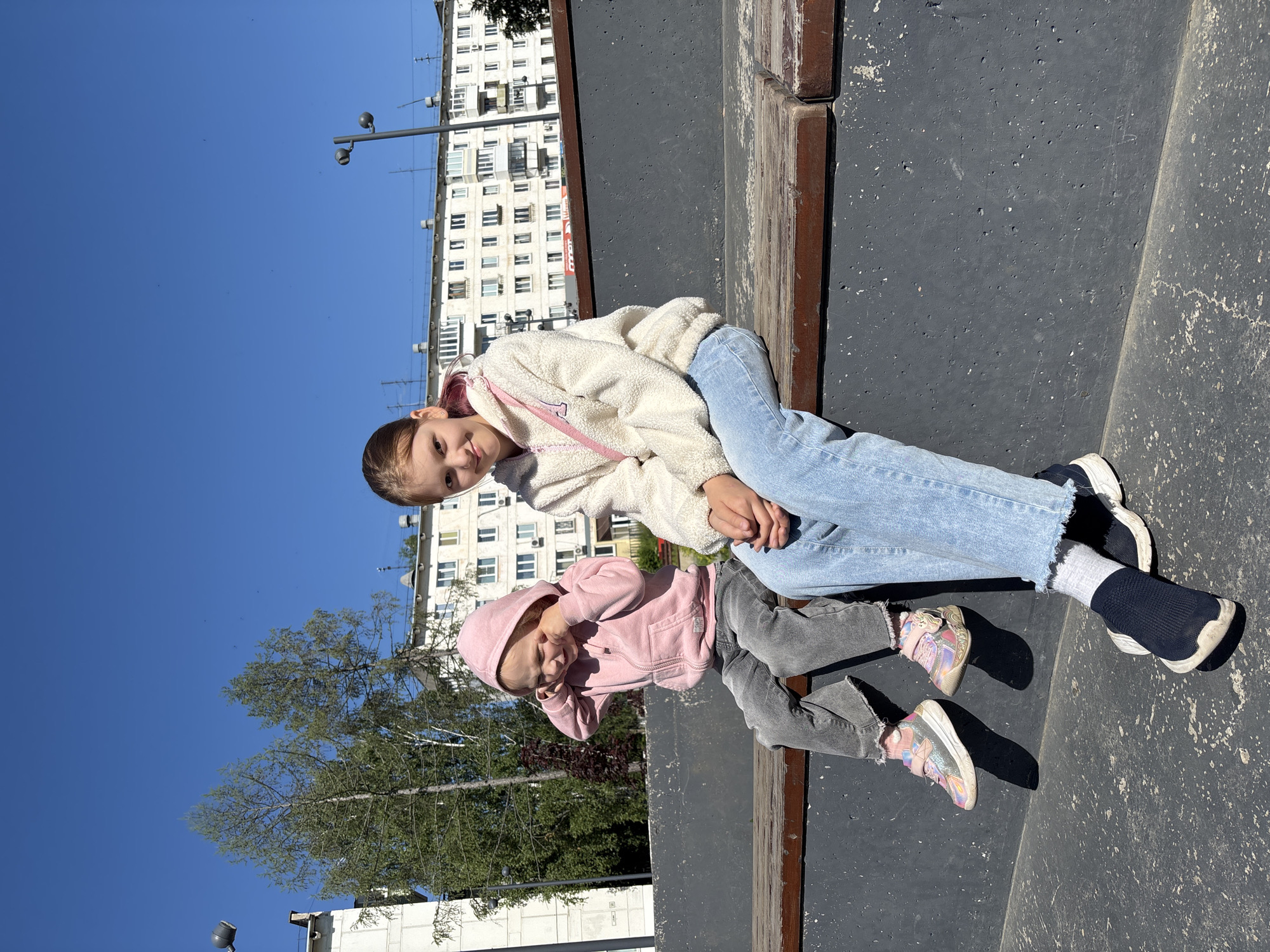 In this outdoor photograph taken in Chaikovsky, Russia, two young girls are seated on a dark, textured surface, likely a skatepark or playground, under a clear blue sky. Sunlight suggests it is daytime.
The older girl, positioned at the top of the frame, wears a fluffy cream-colored hoodie and light blue jeans. She looks towards the camera with a slight smile. A pink strap, possibly from a purse or bag, is visible across her chest.
The younger girl, in the foreground, is dressed in a pink hooded jacket and grey pants. She is leaning forward, with her hands near her face, appearing to be playing or covering her eyes. Her gaze is directed downwards. She wears sparkly pink and silver sneakers. The older girl is wearing dark socks and black and white sneakers.
In the background, a multi-story white building with numerous windows is visible, along with trees and a streetlamp. The overall mood is casual and playful, captured on a bright, sunny day. There is no visible text in the image.