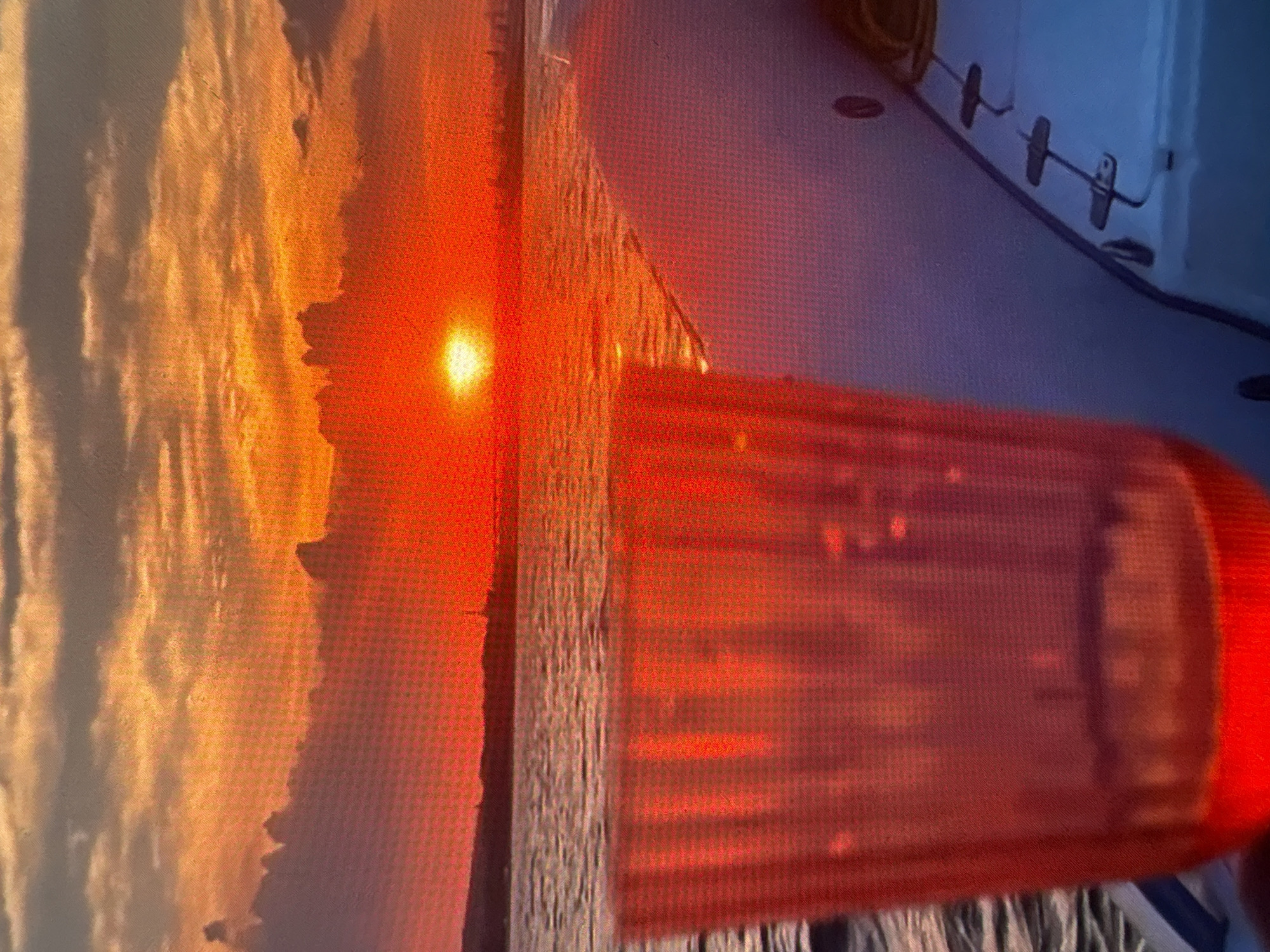 The image shows a sunset view through a red emergency light bar. The sunset displays orange and yellow hues in the sky, with a bright sun visible near the horizon. A dark vertical object, potentially a tree or building, is partially silhouetted against the sunset. The emergency light bar is predominantly red and rectangular, with a blurred appearance. The background includes a section of a room with light blue walls and some fixtures. The location is unknown.