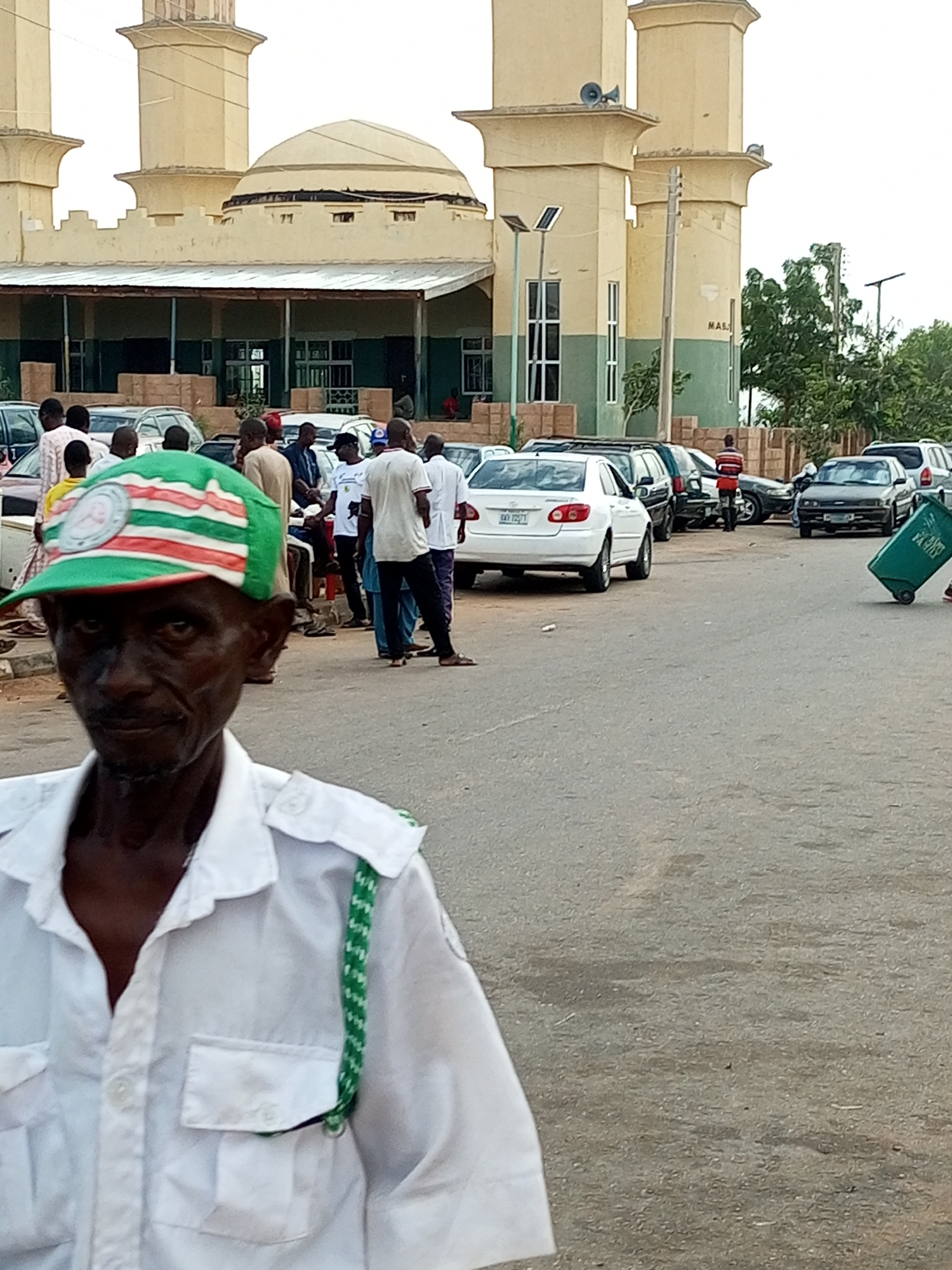 The image depicts an outdoor scene in what appears to be Gombe, Nigeria. In the foreground, a man in a white uniform shirt and a green and white striped cap is looking directly at the camera. He has a focused expression. Behind him, a group of people are gathered near several parked cars.
In the background, a large building with distinctive minarets and a dome, likely a mosque, is visible. The architecture suggests a North African or Middle Eastern influence, common in parts of Nigeria. The sky is overcast, indicating potentially cloudy or subdued lighting, suggesting it might be daytime but not necessarily bright sunlight.
Cars are parked in rows, implying a gathering or event is taking place, or it's a typical scene at a public place like a mosque. Some individuals are standing around, talking or observing. A person is seen pushing a green wheeled bin in the mid-ground to the right, suggesting a cleanup or waste management activity is also occurring.
There is some visible text on a sign on the building in the background, which appears to say "MAS" followed by another partial word. The overall atmosphere seems somewhat busy, with people congregating in front of the prominent building. The presence of multiple vehicles and people suggests a community event or a regular gathering.