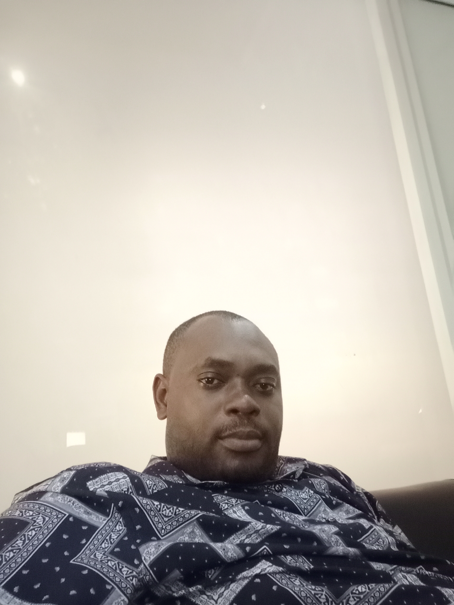 A medium-shot photograph depicts a dark-skinned man in his late 30s or early 40s. He has short, dark hair, and a mustache. The man is wearing a button-down shirt with a dark blue pattern. He appears to be sitting on a dark-colored couch or chair. The background is a plain, off-white wall with no distinguishing features. The location is Gwarinpa, Nigeria, as requested. There are no actions or interactions visible, and the environment appears to be indoors.