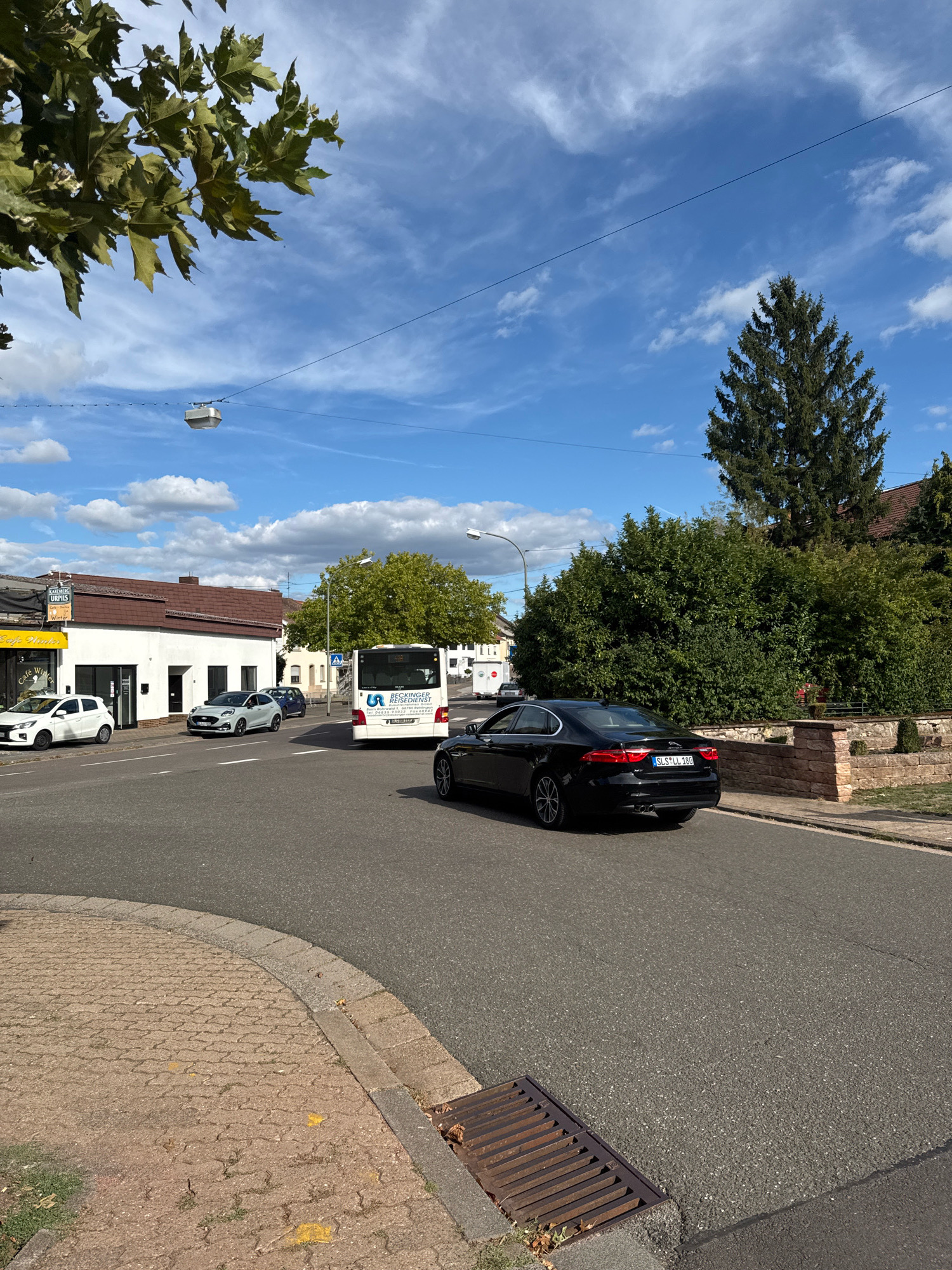 This is a street-level, medium-shot photograph taken in Überherrn, Germany, under a partly cloudy blue sky on a sunny day. The image captures a road intersection with several vehicles and buildings. In the foreground, a black sedan, identified by its license plate "SLS LL 180", is moving forward on the asphalt road, which has a distinct cobblestone curb and a metal grate drain on the right. To the left, a white hatchback is parked in front of a white building with a red-tiled roof. Next to it, a light gray hatchback is also parked. Further down the street, a white bus with "BECKINGE REISEDIENST" text on its rear is traveling away from the camera. The background features a dense green hedge and a tall evergreen tree on the right, with residential buildings visible behind the foliage. On the left side of the road, a yellow awning with text including "Cafe Winter" is attached to a building. Overhead, power lines with small light fixtures are visible.