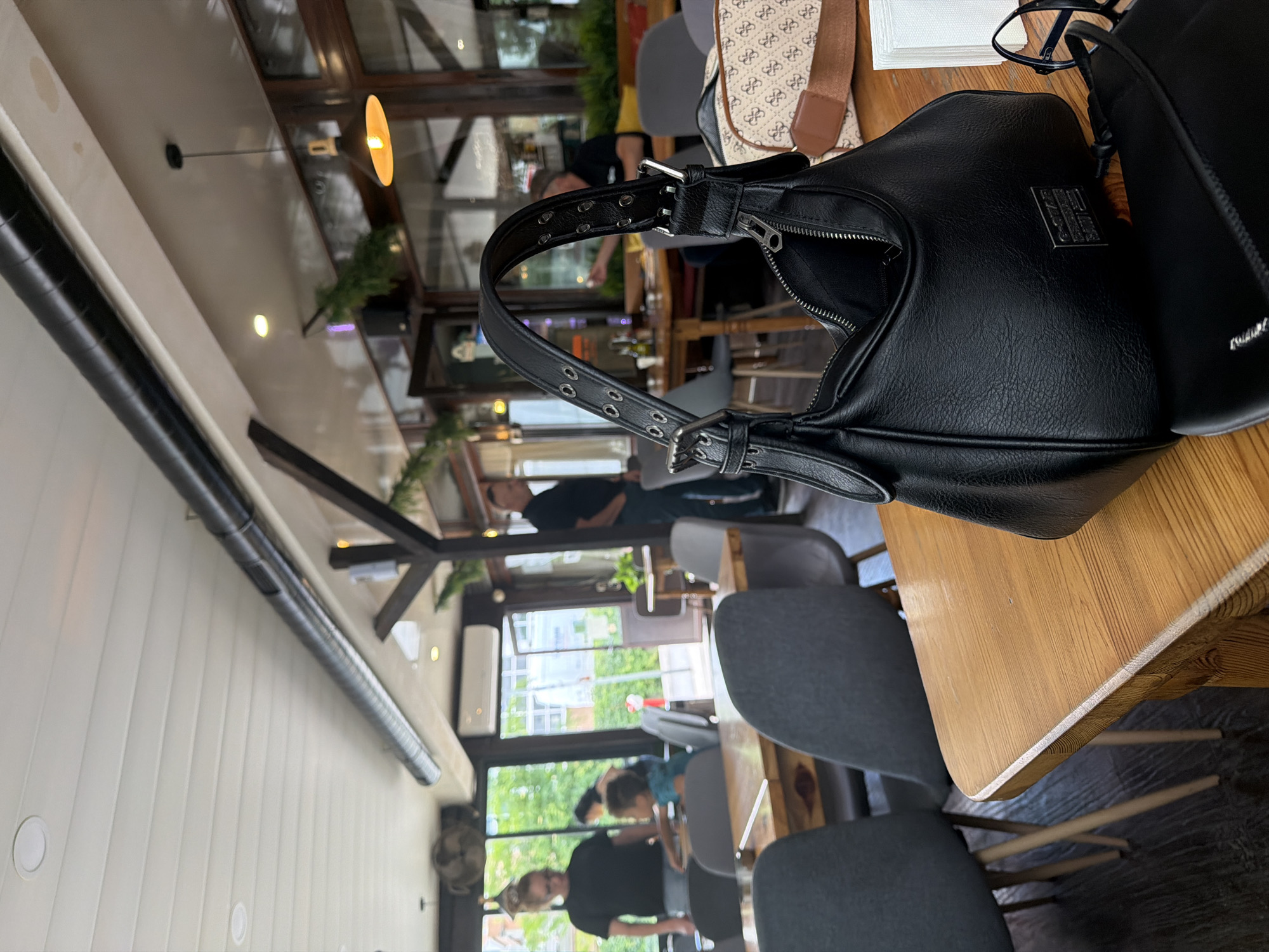 This is a medium shot photo, taken indoors at a restaurant or cafe, with people seated at tables. The photo is taken at a slightly elevated angle, looking down towards the table and chairs. The primary focus is a black leather handbag with silver buckle details on the table, along with another bag, and a pair of glasses. In the background, through large windows, lush green trees are visible, suggesting a pleasant outdoor environment. The lighting suggests it might be daytime, possibly afternoon. The scene appears relaxed and casual, with individuals engaged in dining or conversation. The image captures a slice of everyday life in a cafe setting. No visible text is present in the image.