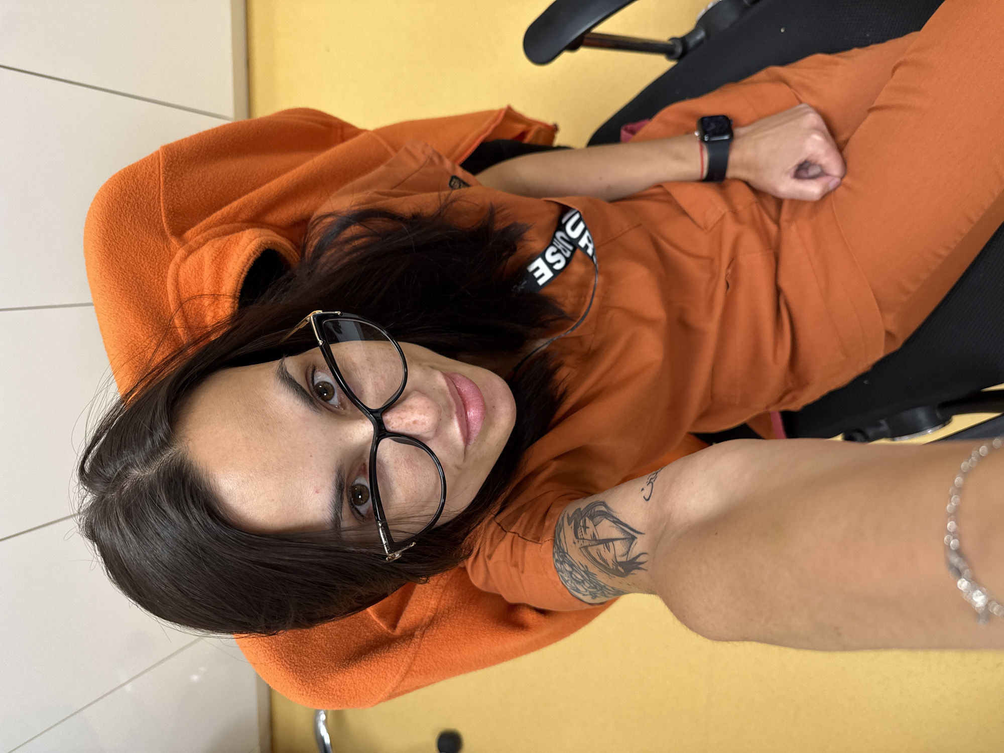 A woman with dark hair and glasses is sitting in a black office chair. She is wearing an orange hoodie and matching pants, with a black smartwatch on her left wrist. A tattoo of a ship is visible on her left forearm, and a silver bracelet is on her right wrist. The background consists of a yellow floor and a white wall with horizontal lines. The photograph appears to be taken indoors, possibly in an office setting, and is a close-up, high-angle shot. There is no indication of this being taken in Pleven, Bulgaria.