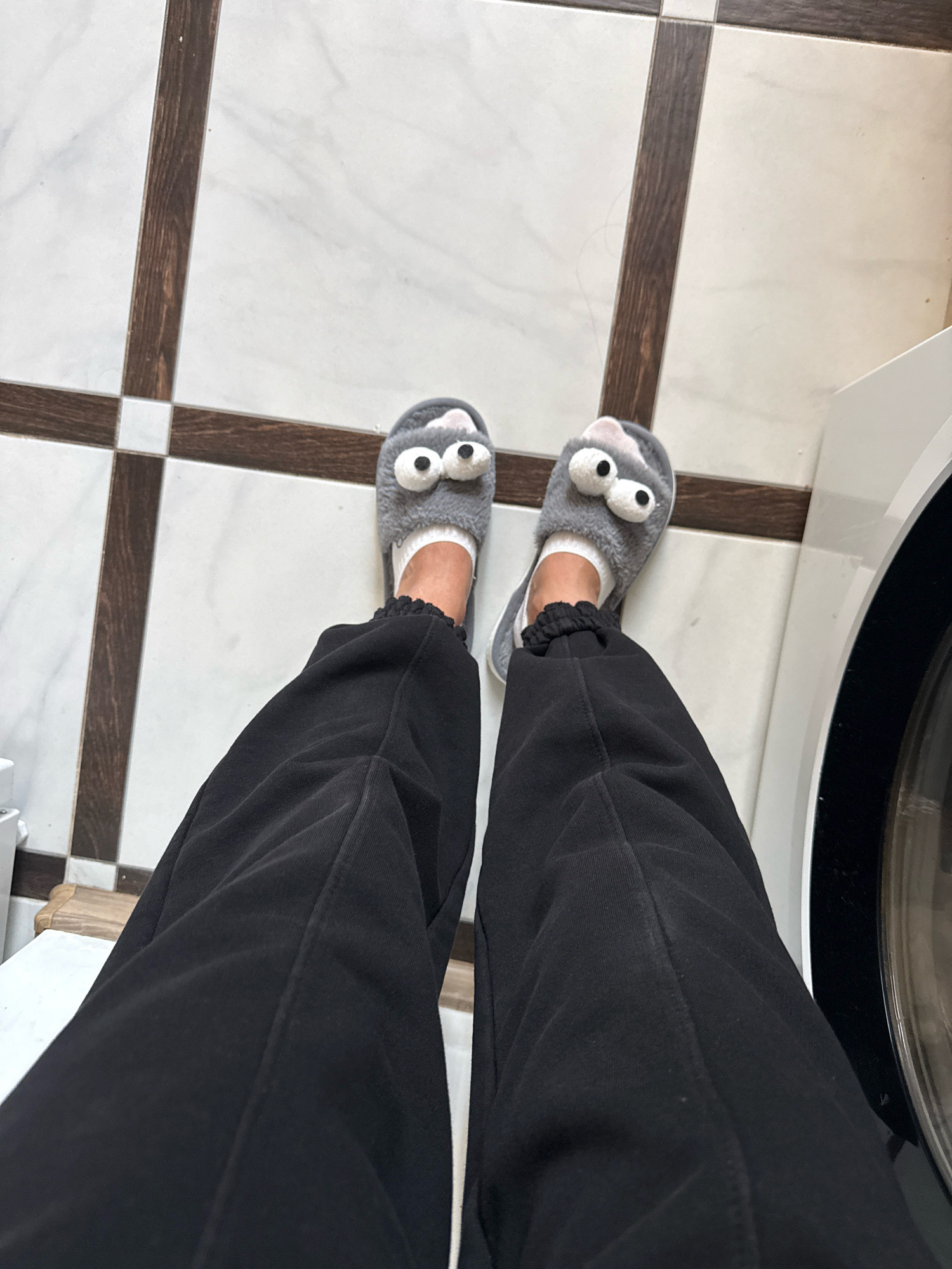 The image shows a close-up, overhead view of a person's feet wearing fuzzy grey slippers with googly eyes and white socks. The slippers are on a tiled floor with white tiles and dark brown grout lines. The person is wearing black sweatpants. To the right of the feet, a portion of a white appliance, possibly a washing machine, is visible. The scene appears to be indoors, likely in a residential setting. The location is not specified in the image, and therefore cannot be identified as being in Chișinău, Moldova.