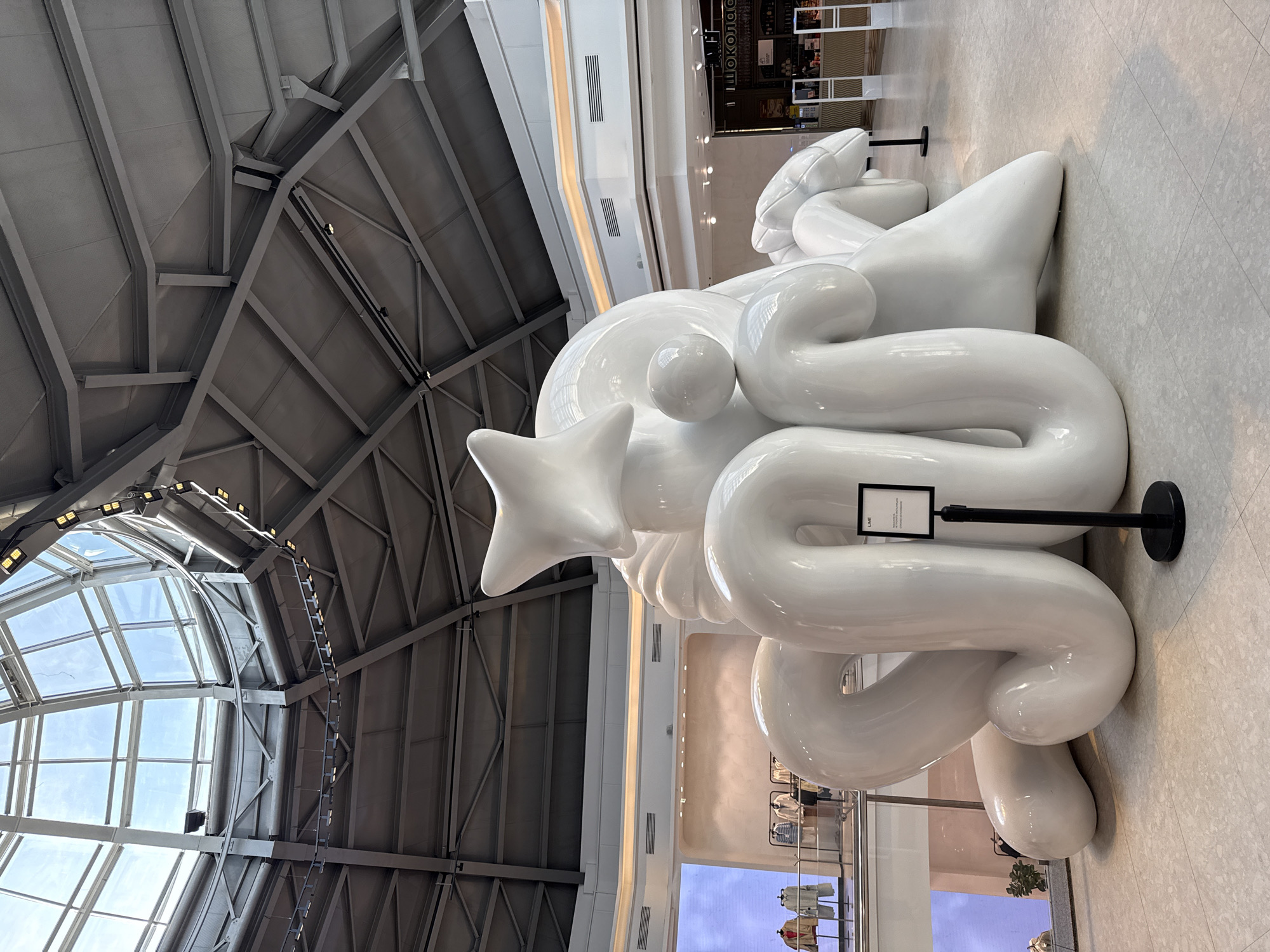 The image shows a large, white, abstract sculpture in a modern, indoor space, likely a shopping mall. The sculpture is composed of smooth, rounded shapes, including a star. It is roped off by black stanchions and a sign is posted next to it. The mall has a high ceiling with a combination of metal beams and a glass skylight. The floor is a light-colored tile. In the background, various retail stores are visible. The sculpture is located in the Aviapark shopping mall in Moscow, Russia.