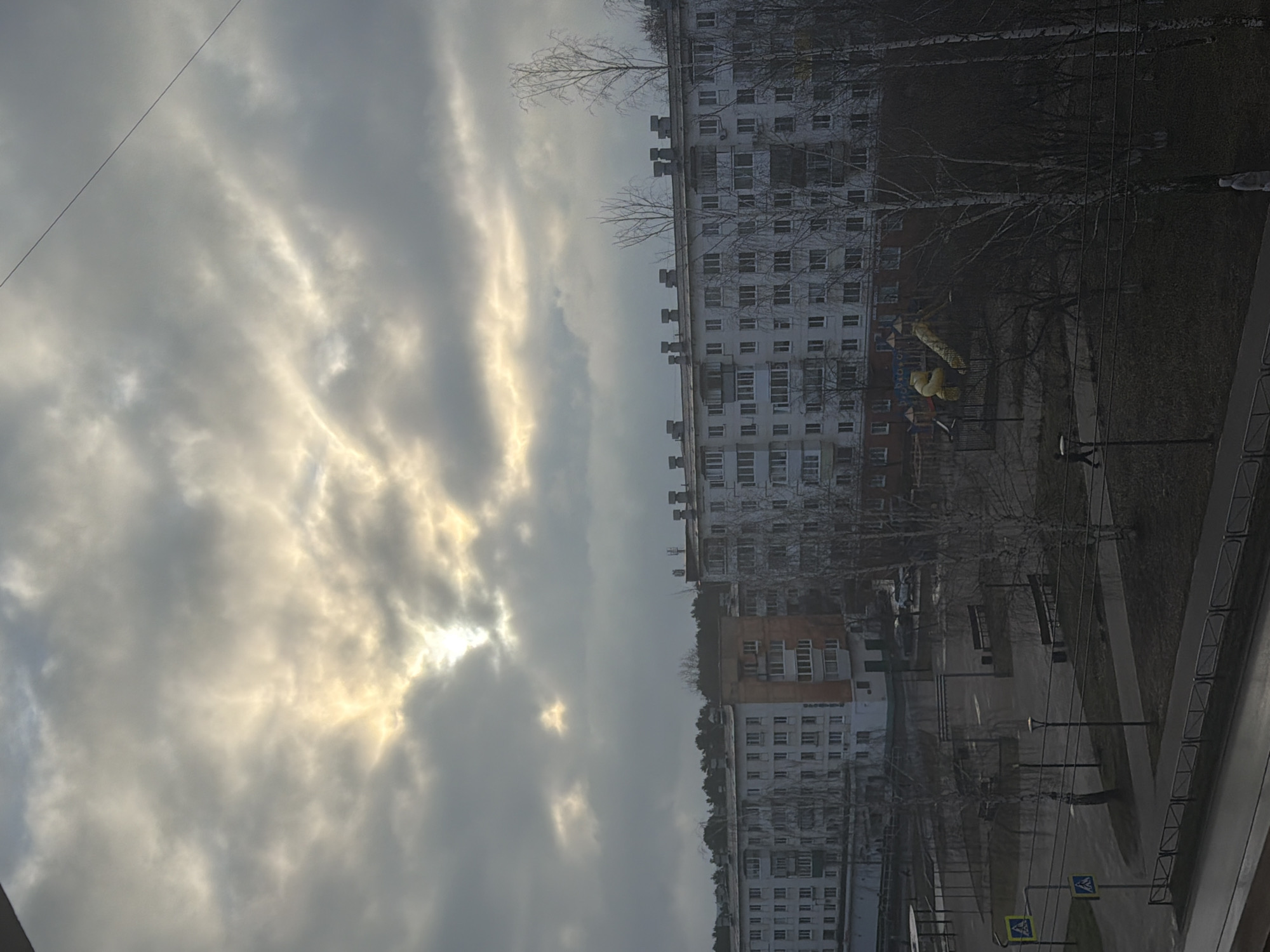 The image is a ground-level, outdoor shot in the city of Chaikovsky, Russia. The view is from an elevated position, possibly a window. The sky is overcast with heavy, streaky clouds, with the sun trying to break through. Below, there are several high-rise apartment buildings, constructed with many windows on each floor. There is also a playground with yellow and blue structures visible. A grassy area interspersed with bare trees and walkways can be seen. Sidewalks with railings are in the foreground, suggesting a pedestrian area. There are also street lamps and zebra crossing signs on the road. The overall environment seems quiet. No people can be clearly identified due to the distance and quality of the image.