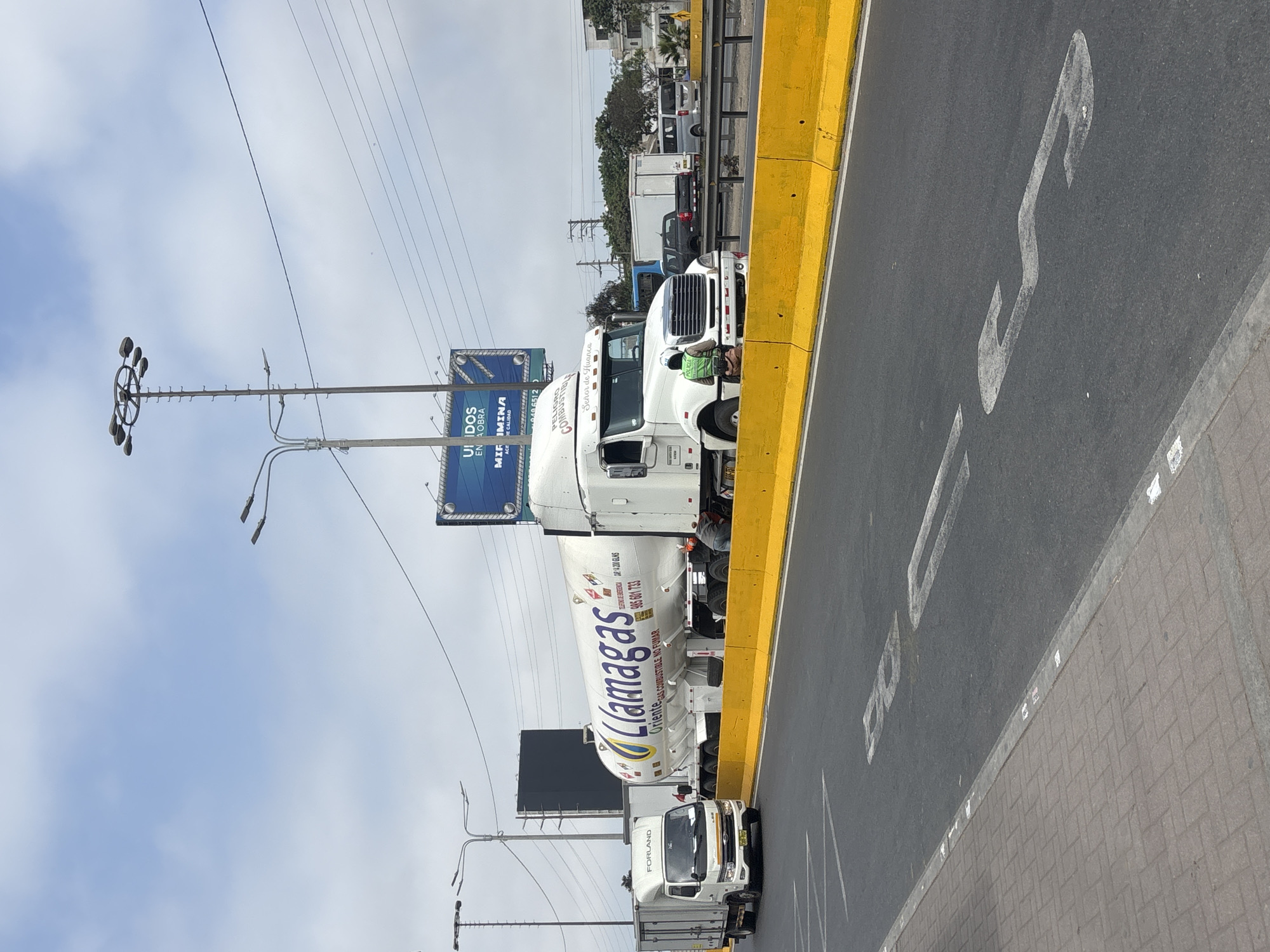 The image captures a busy roadside scene in San Juan de Miraflores, Peru, on a clear, cloudy day. Several trucks are visible on a paved road next to a yellow barrier.
In the foreground, a large white tanker truck belonging to "Llamagas" is prominently featured. The side of the tank reads "Llamagas," "Oriente," "GAS COMBUSTIBLE NO FUMAR" (Combustible Gas Do Not Smoke), "TACAR 14 200 GLNS," and the phone number "985 601 733." Directly in front of the tanker, a white tractor-trailer is parked, with "PELIGRO COMBUSTIBLE" (Danger Combustible), "Señor de Huanca," and "049 651 2" visible on its side. A person wearing a bright green safety vest is seen near the front wheel of this tractor-trailer, seemingly inspecting something or working. Further back, a smaller white cargo truck with the brand "FORLAND" on its cab is also present, along with other vehicles in the background.
The road features white painted markings, including what appears to be a large "S" or serpentine shape. Adjacent to the road is a sidewalk made of grey pavers. Overhead, power lines crisscross the sky, and multiple streetlights are mounted on poles. In the mid-ground, a large blue billboard displays text including "UNIDOS EN OBRA" (United in Work), "MIR IMINA," and "DE CALIDAD." The scene depicts typical urban or highway infrastructure, with vehicles potentially in transit or stopped for maintenance.