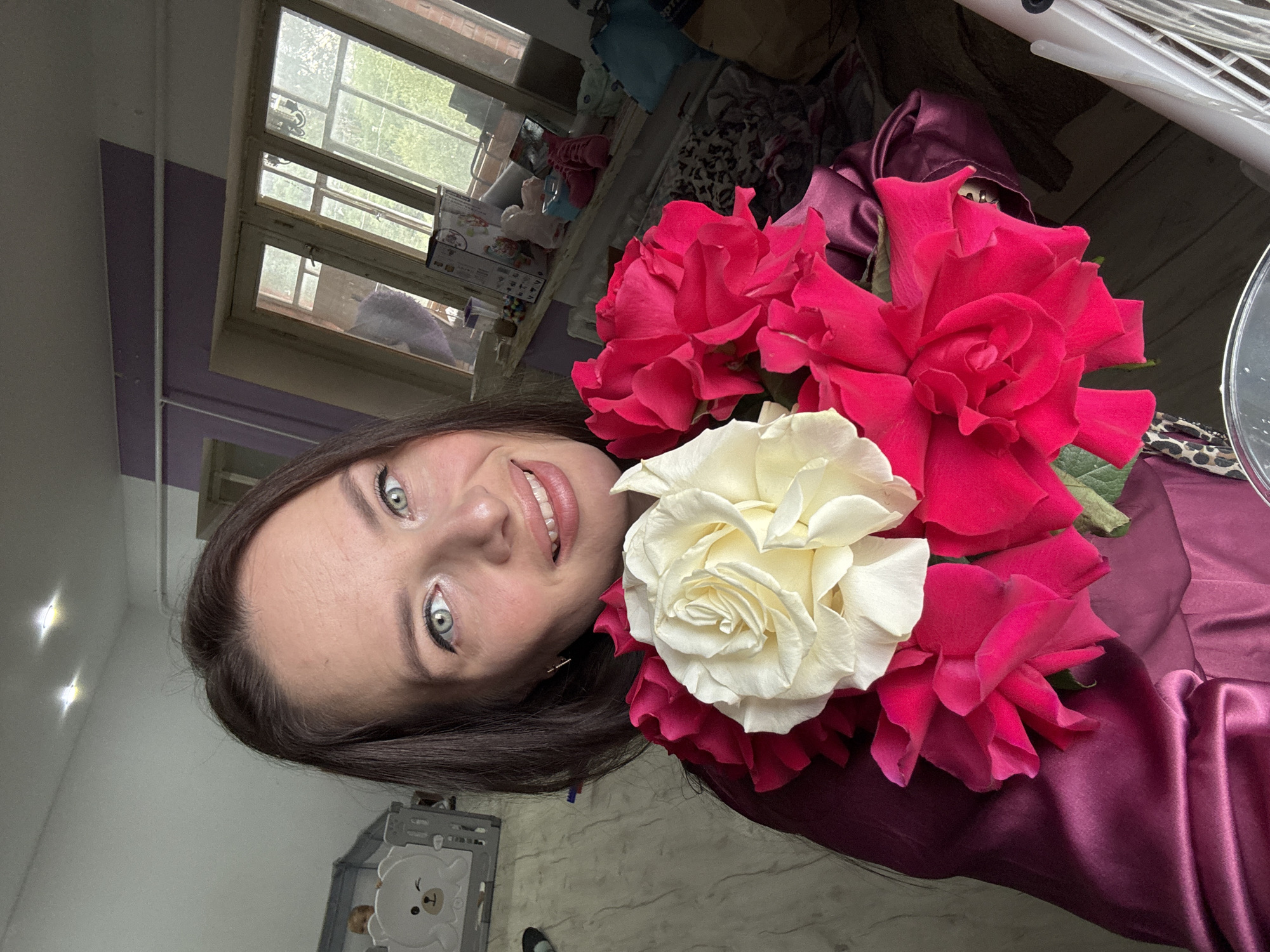 A woman with brown hair and blue eyes is holding a bouquet of roses, with one white rose and several pink roses. She is smiling at the camera. She is wearing a dark pink satin dress. In the background, there is a baby crib with a teddy bear design on it, a window with sunlight streaming through, and various objects on a windowsill. The location appears to be an indoor residential setting.