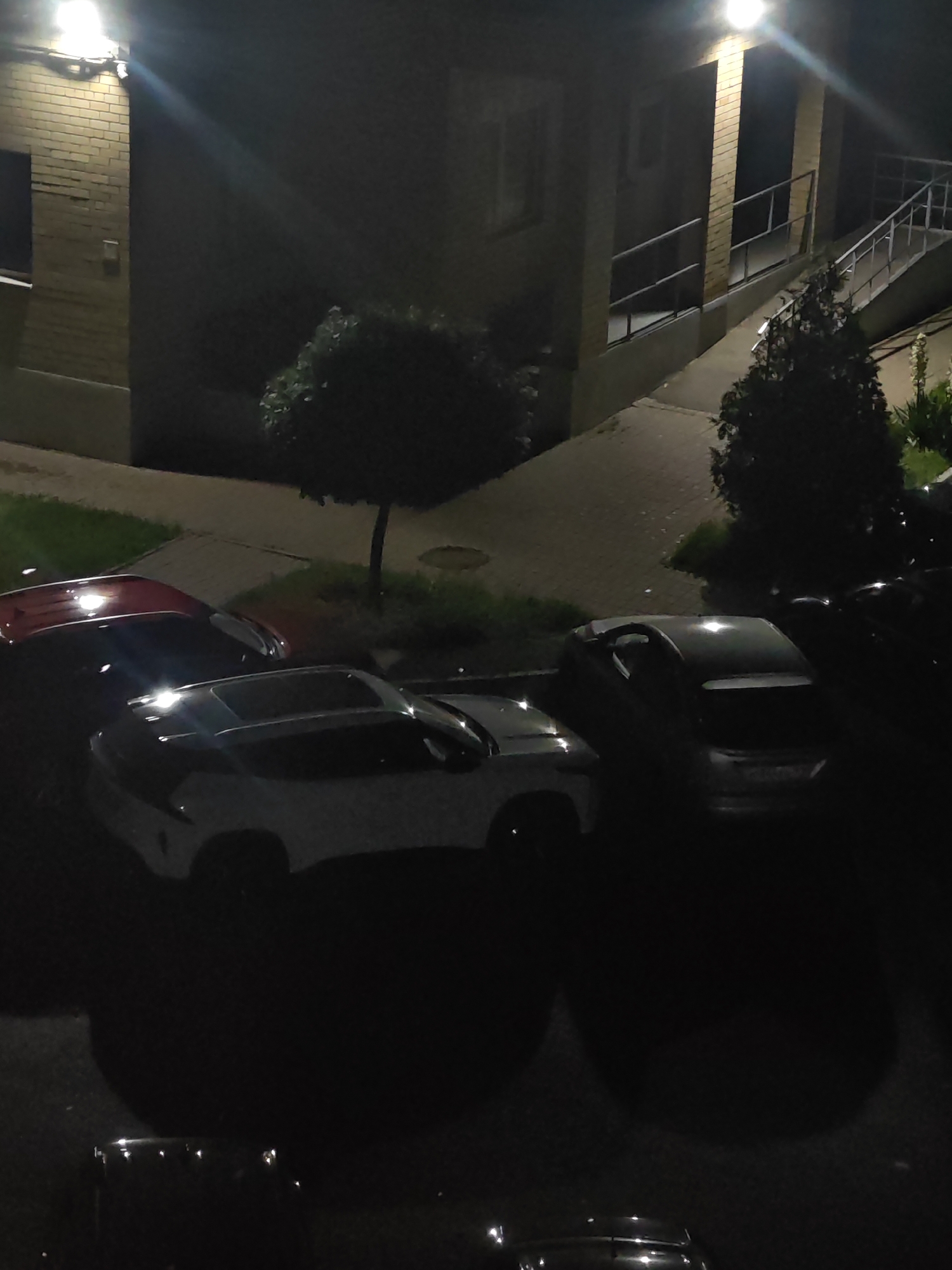 The image depicts a nighttime scene in an outdoor urban environment, likely a residential area or a parking lot. A brick building with a light fixture illuminating the facade is visible on the left. A paved walkway with a railing leads up to the building, and a ramp is also present. Several cars are parked in the foreground, including a red car, a white car, and a darker colored car. The lighting is dim, with the primary illumination coming from the building's light fixture and reflections on the cars. The presence of trees and grass suggests a natural element within the urban setting. The image is devoid of any human presence. There is no information provided about the specific location within Bataysk, Russia.