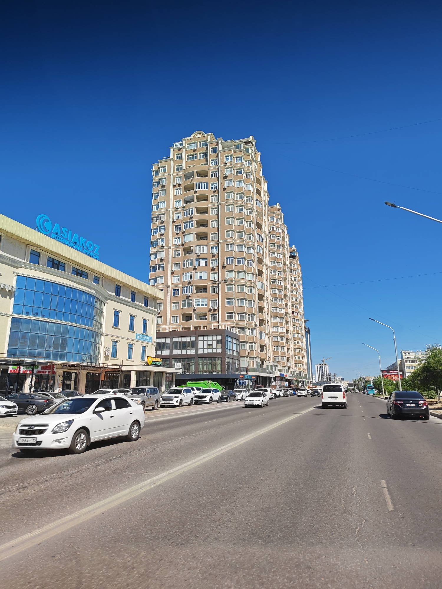 This is a street-level, wide-angle, eye-level, full shot of a city street in Aktau, Kazakhstan, on a clear, sunny day. The street is paved and appears to be a multi-lane road. In the foreground, the road surface shows some wear and tear. Several cars are visible on the road, including a white Chevrolet sedan in the left lane, and a white van in the right lane further down the street. A dark sedan is visible in the right lane at the far end of the image.
Along the left side of the street, a modern, multi-story building with a light-colored facade and blue glass windows dominates the scene. A prominent blue sign with the text "ASIAKOZ" is displayed at the top of this building, indicating it might be a business or commercial establishment. Adjacent to this building, and slightly behind it, are tall, beige apartment buildings with numerous balconies and windows. The architecture of these buildings suggests a residential area.
On the right side of the street, trees and other buildings are visible in the distance. Streetlights are present along the right side of the road. The sky is a vibrant, clear blue, with no clouds visible. The overall impression is a bustling urban environment in Aktau.