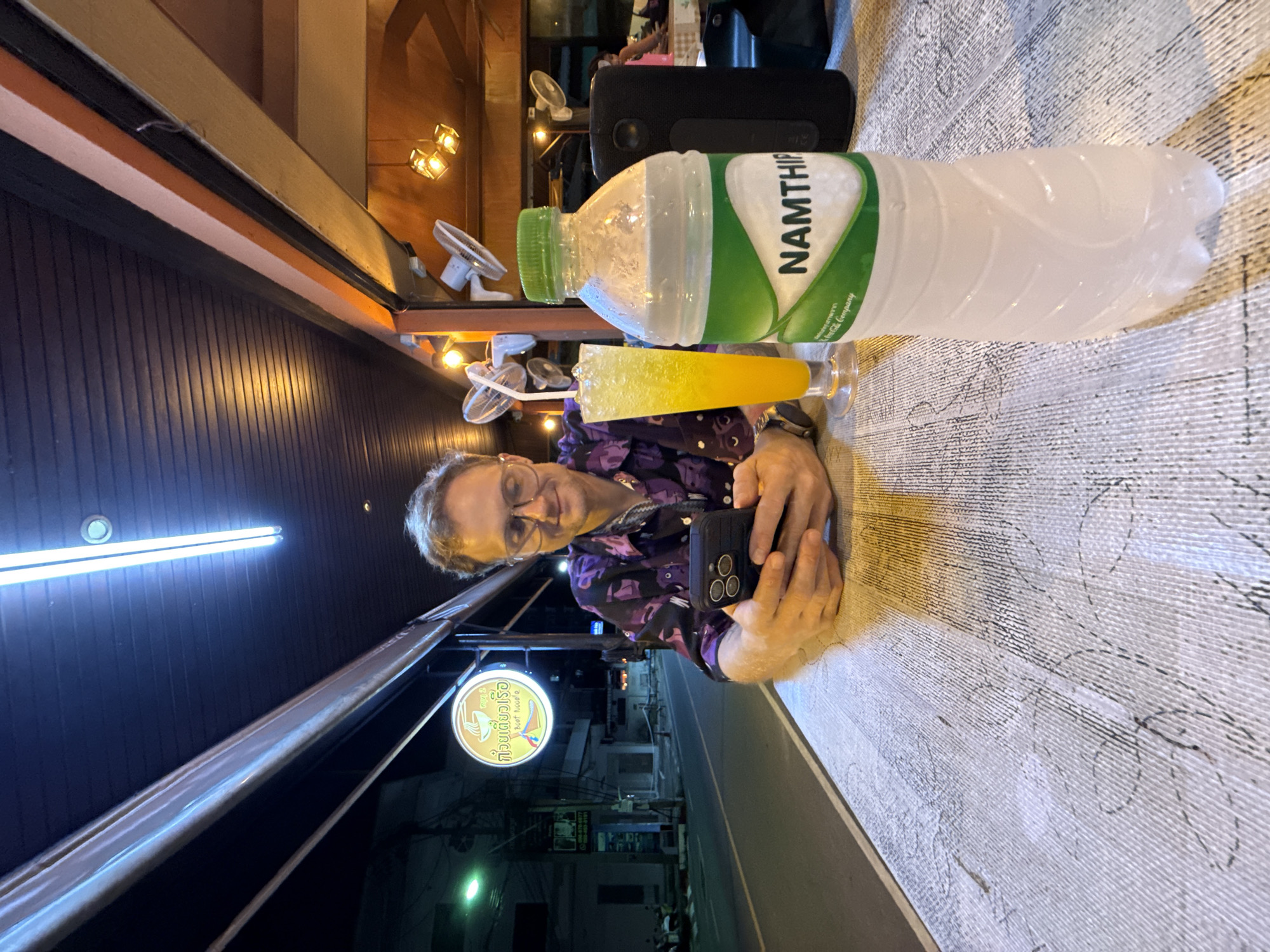 In this close-up, medium shot, a Caucasian man with glasses and curly hair sits at a table outdoors at night. He is wearing a purple patterned shirt and looking down at his phone, which he is holding in his hands. In front of him is a glass of yellow liquid with ice and a straw, and a bottle of Namthip water. The table has a textured surface with a pattern that resembles wood. Behind the man, a street is visible with buildings and a neon sign with Thai writing. The lighting is dim, with overhead lights casting a glow on the scene.