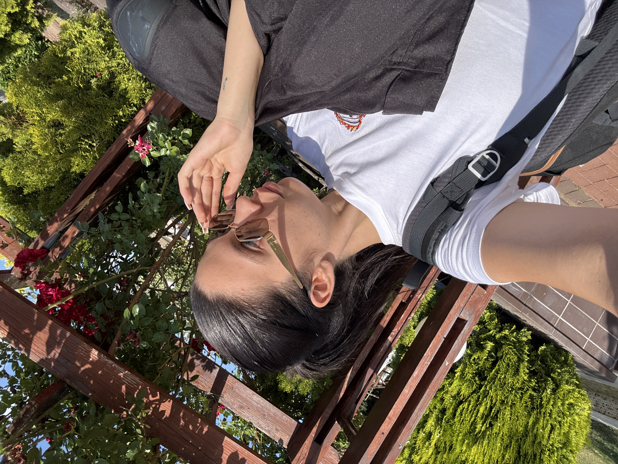 A young woman with dark hair tied in a ponytail, wearing sunglasses, a white t-shirt, and dark pants, is pictured outdoors. She is leaning back, with her hand near her face, and appears to be sitting on a bench. She is wearing a backpack. The background consists of a wooden pergola with red roses and green bushes. The scene suggests a sunny day, possibly in the city of Chavdar, Bulgaria.