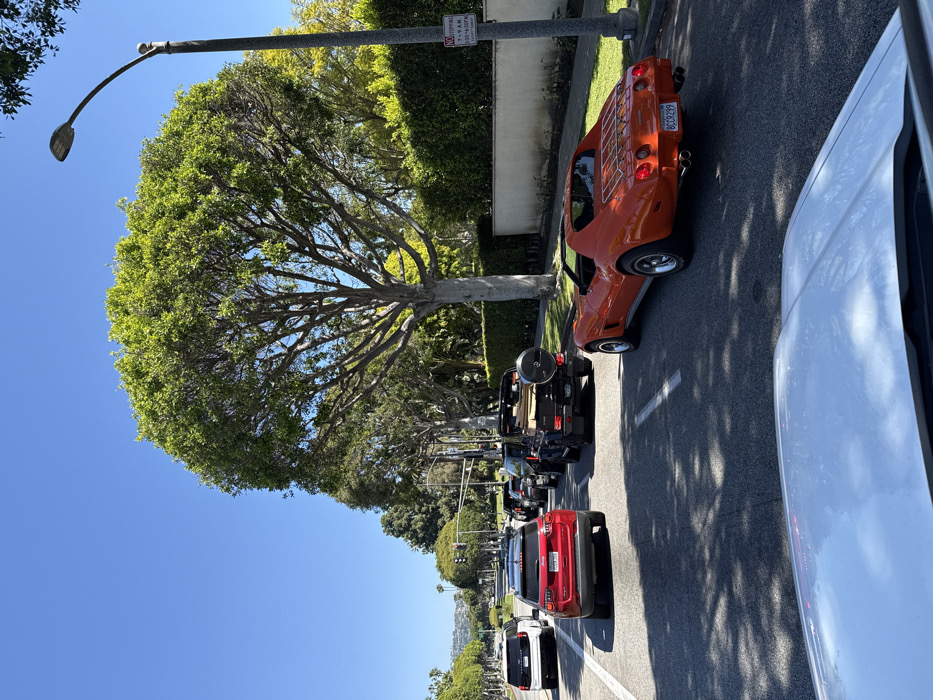 The image depicts a street scene under a clear blue sky on a sunny day in Beverly Hills, United States of America, observed from the interior of a vehicle. Traffic is present in multiple lanes, appearing to be stopped or moving slowly.
In the immediate foreground, the hood and windshield of a light-colored vehicle are visible. Ahead in the right lane, an orange Chevrolet Corvette, likely a C3 generation with a t-top configuration (tops removed), is positioned. This vehicle features a chrome luggage rack on its rear deck lid and dual exhaust pipes on each side. A California license plate, "8CCR299," is affixed to its rear.
Behind the Corvette, in the adjacent lane, a black Mercedes-Benz G-Wagon is visible, equipped with an external spare tire mounted on its rear door. Further back in the same lane, a red Jeep SUV is present. In the leftmost lane, a white Range Rover SUV is visible, followed by several other vehicles receding into the distance.
The street is paved with asphalt and marked with white lane lines. On the right side of the road, a wide, mature tree with dense green foliage casts shadows onto the pavement. Adjacent to this tree, a grey street lamp with an LED fixture is installed. A white regulatory sign, indicating "NO STOPPING 7-9 AM EXCEPT SUNDAY & HOLIDAYS" within a red circle and diagonal line, is attached to the streetlight pole. Behind the tree and pole, a well-maintained dark green hedge runs along a light-colored wall, demarcating residential property. In the distance, low hills or mountains are visible against the horizon. No individuals are clearly discernible.