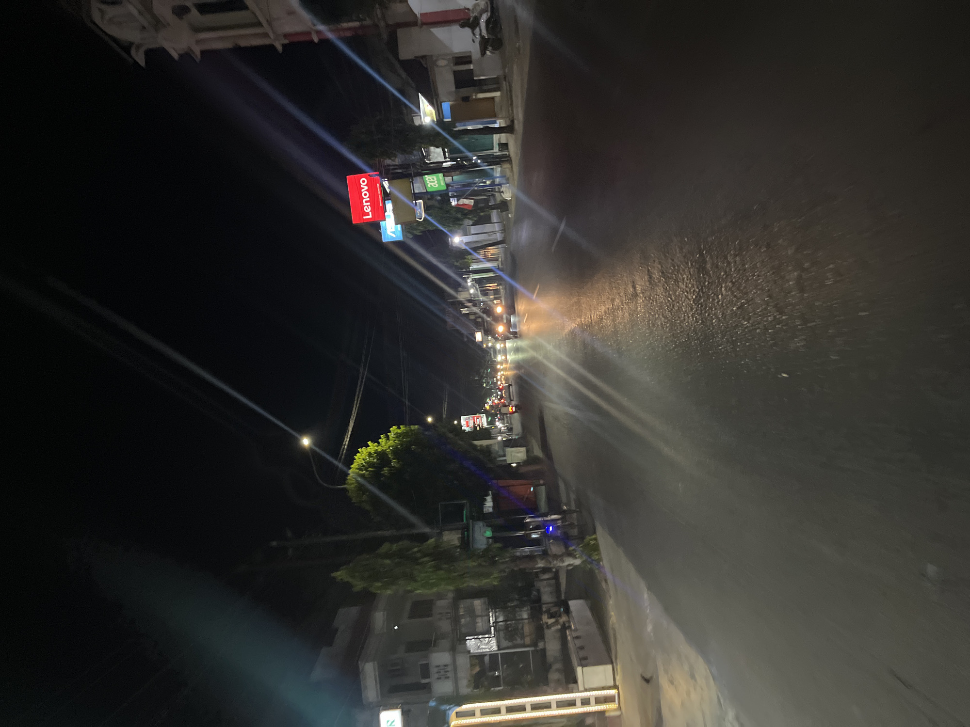 This is a night-time street scene in Kalinyamatan, Indonesia. The asphalt road is wet and reflects the light from street lamps and vehicle headlights, creating streaks of light across the image. On the left side of the road, there are buildings with multiple stories, some with windows and balconies. One building has a vertical illuminated structure. Trees are visible along the roadside, with one tall, slender tree in the foreground. In the distance, traffic lights and the headlights of approaching vehicles can be seen. A prominent red sign with the "Lenovo" logo is visible on the right side of the street, mounted on a pole. Next to it, another sign with "acer" in green lettering is also present. A few motorcycles are parked or moving on the road, and a person is visible on a motorcycle in the far right of the image. The sky is dark, indicating it is nighttime.