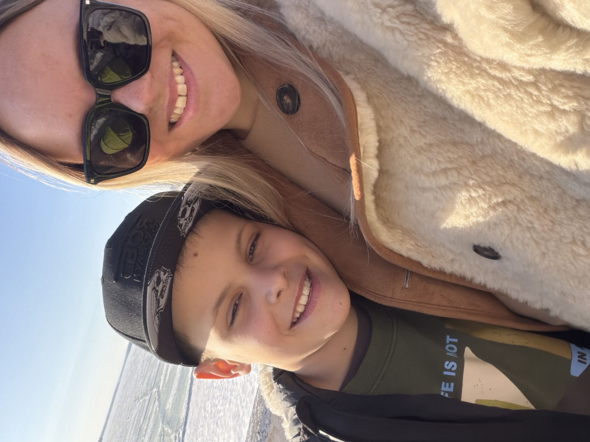 Two people are posing for a selfie. A woman with blonde hair is wearing sunglasses and a tan coat with a light-colored fur lining. A boy is wearing a black baseball cap and a green sweatshirt with the partial words "LIFE IS NOT" visible. They are both smiling. The background appears to be a bright, outdoor setting. The specific location in Votkinsk, Russia, cannot be determined from the visual information provided.