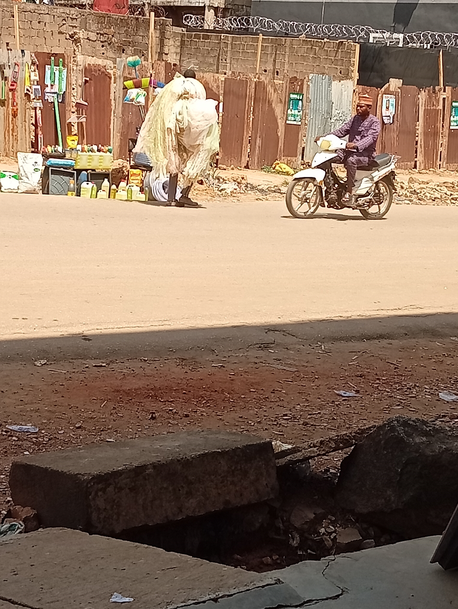 Maiburiji, Nigeria. Daytime. Mid-shot of a street. Foreground shows broken concrete and dirt. Mid-ground shows a street where a vendor with household supplies is standing. Person appears to be loading something onto their back. Further along the street, a man is riding a white motorcycle. Background shows fencing and buildings.