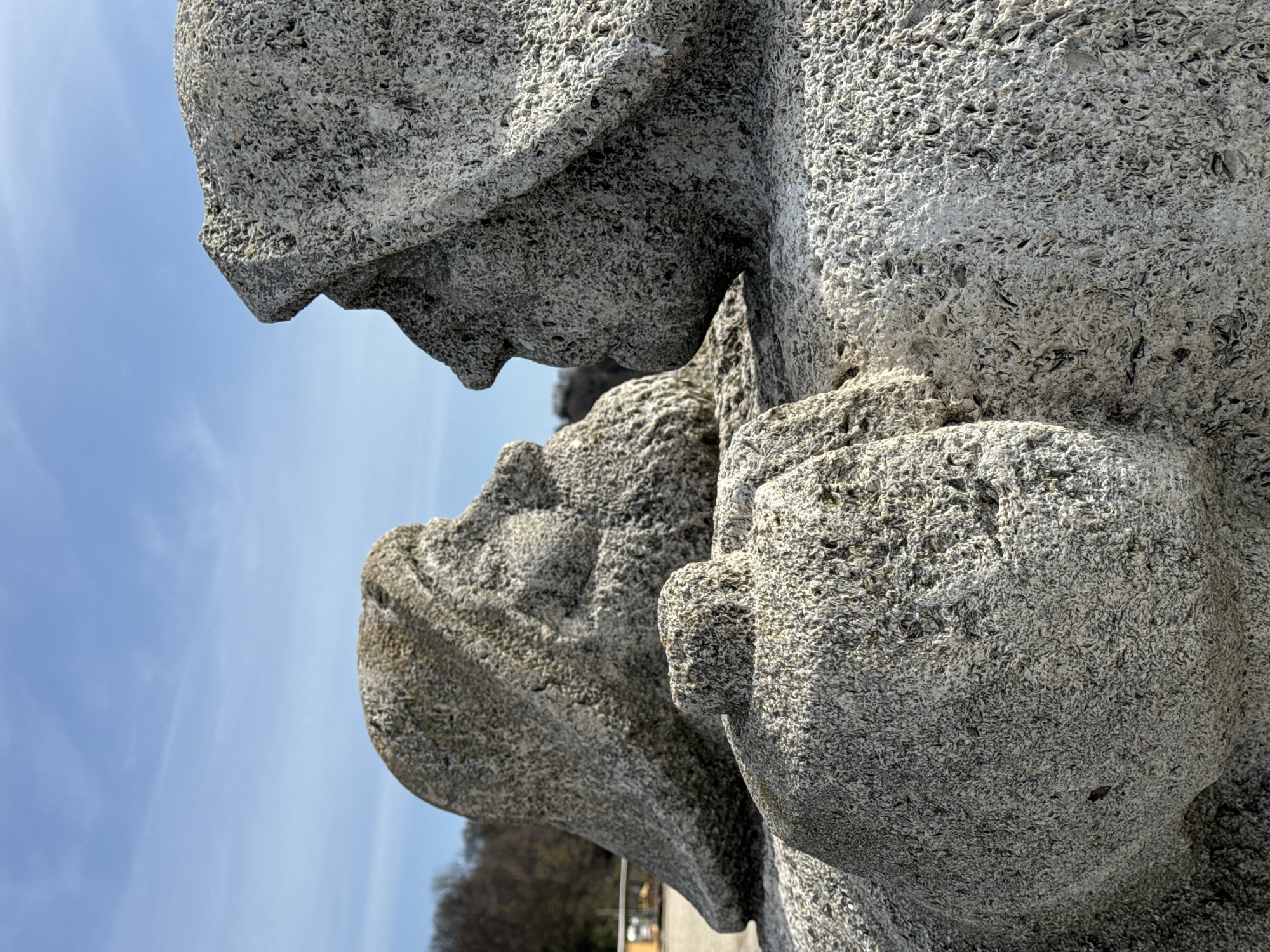 A low-angle, close-up shot presents a row of three concrete, head-and-shoulder busts of individuals, possibly military figures. The busts are lined up one behind the other. The sky is visible to the side. The scene takes place in Varna, Bulgaria. The surface texture of the concrete is rough and pitted, with an appearance suggestive of limestone. In the background, there are trees and a building.
