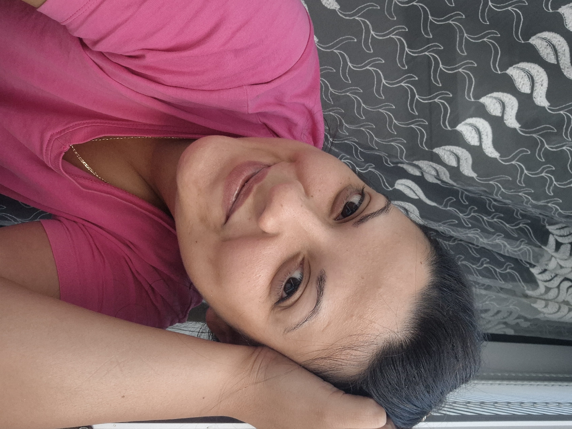 In a selfie taken indoors, a woman with dark hair and brown eyes smiles as she rests her head on her hand. She is wearing a bright pink t-shirt and a delicate gold chain necklace. The background features a grey curtain with a white leaf-like pattern, and a portion of a window frame is visible on the right. The overall impression is one of relaxed contentment. The image was taken in Lovech, Bulgaria.
