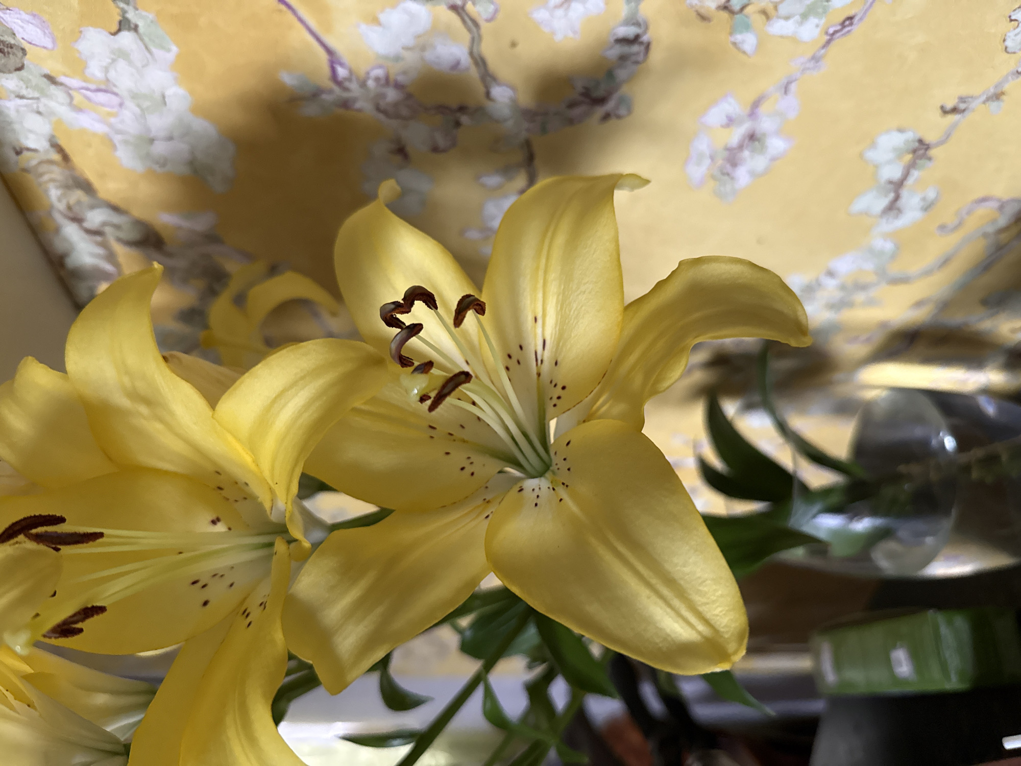 A close-up shot shows two yellow lilies in bloom, with their petals unfurled and their stamens visible. One lily is in the foreground, its petals a vibrant yellow with dark speckles near the center. The other lily is slightly out of focus behind it. The background features a yellow wallpaper with a delicate floral pattern of white and purple blossoms on thin branches, reminiscent of Japanese art. To the left of the lilies, a green bottle and a clear glass vase containing some greenery are partially visible. The lighting is soft, casting a gentle glow on the flowers. The scene suggests an indoor setting, possibly a home or studio, where the lilies are displayed as a decorative element. There are no people or animals present in the image. The location is not specified in the image, but the overall aesthetic suggests a serene and artistic environment.