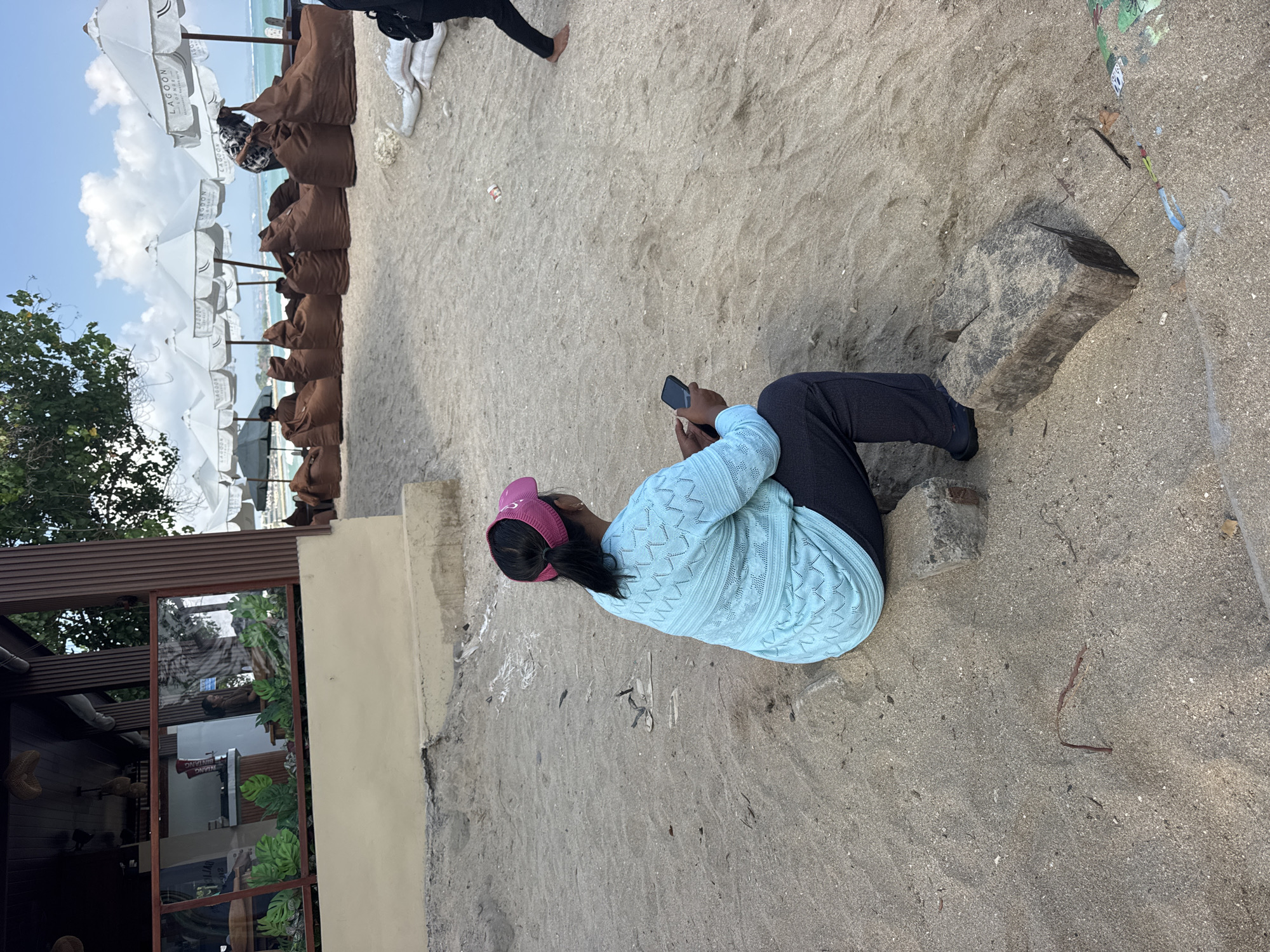 A woman is sitting on the sandy beach in Kuta, Indonesia, holding a smartphone. She is wearing a light blue patterned long-sleeved shirt and dark pants, and a pink visor. She is seated on two concrete blocks. In the background, there are rows of brown beanbag chairs and white umbrellas with "LAGOO..." visible on them. A building with a thatched roof is to the left. The ocean and sky with clouds are visible in the distance.