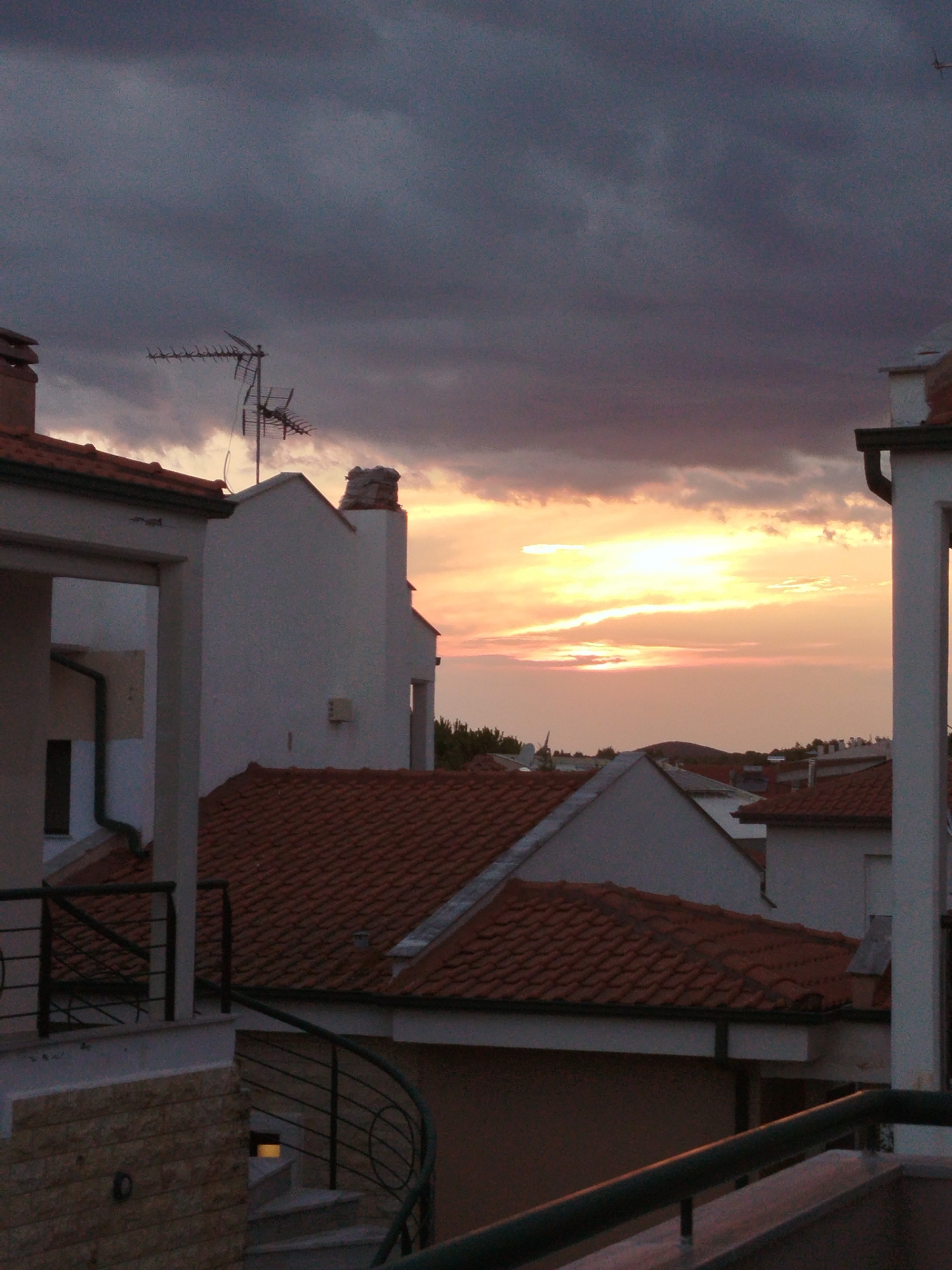 The image captures a dusky scene in Sithonia Municipal Unit, Greece, with a sunset casting a warm glow through dramatic, dark clouds. The foreground is dominated by the terracotta tiled roofs of residential buildings, characteristic of Mediterranean architecture. A white building with a visible drainpipe is positioned to the left, while a taller white structure with a chimney stands to the right. An old-fashioned television antenna is mounted on a rooftop, its metal frame stark against the sky. The overall impression is one of a quiet, late-day residential area, bathed in the fading light of the sun.