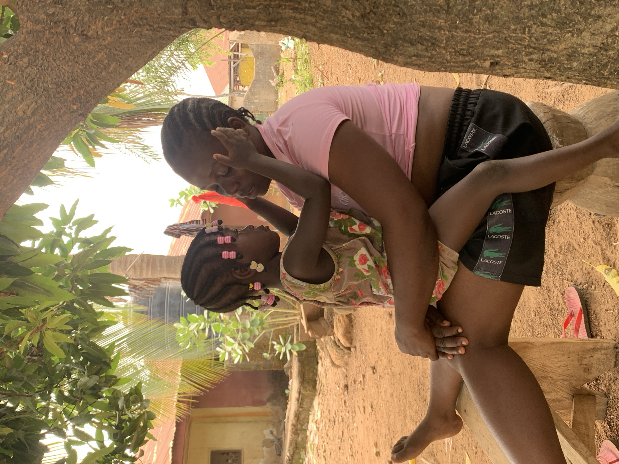 In Akwanga, Nigeria, a young woman in a pink shirt and black Lacoste shorts holds a young girl in a floral dress. The girl has pink and yellow beads in her braided hair. They are facing each other closely, possibly interacting affectionately. They are positioned near a wooden structure, possibly a bench or stool, in an outdoor area with trees, a dirt ground, and glimpses of buildings in the background. A red and black sandal is near the wooden structure.