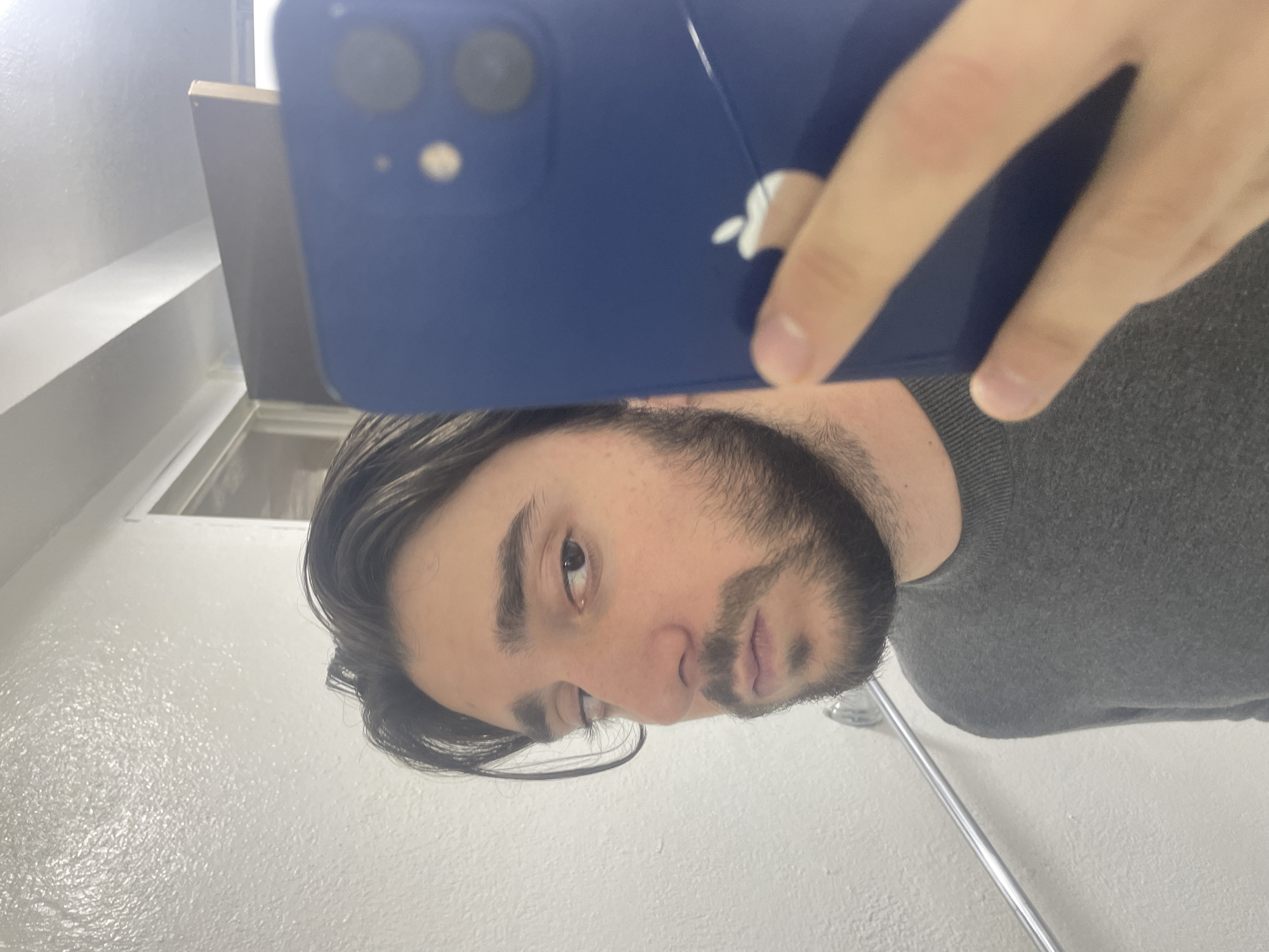 A man is taking a selfie with a blue iPhone in a bathroom. The background consists of a white wall, a portion of a mirror, and a shower curtain rod. The man has dark hair and a beard, and he is wearing a gray sweater. There is no indication of this photo's location being in the city of Huixquilucan de Degollado, Mexico.