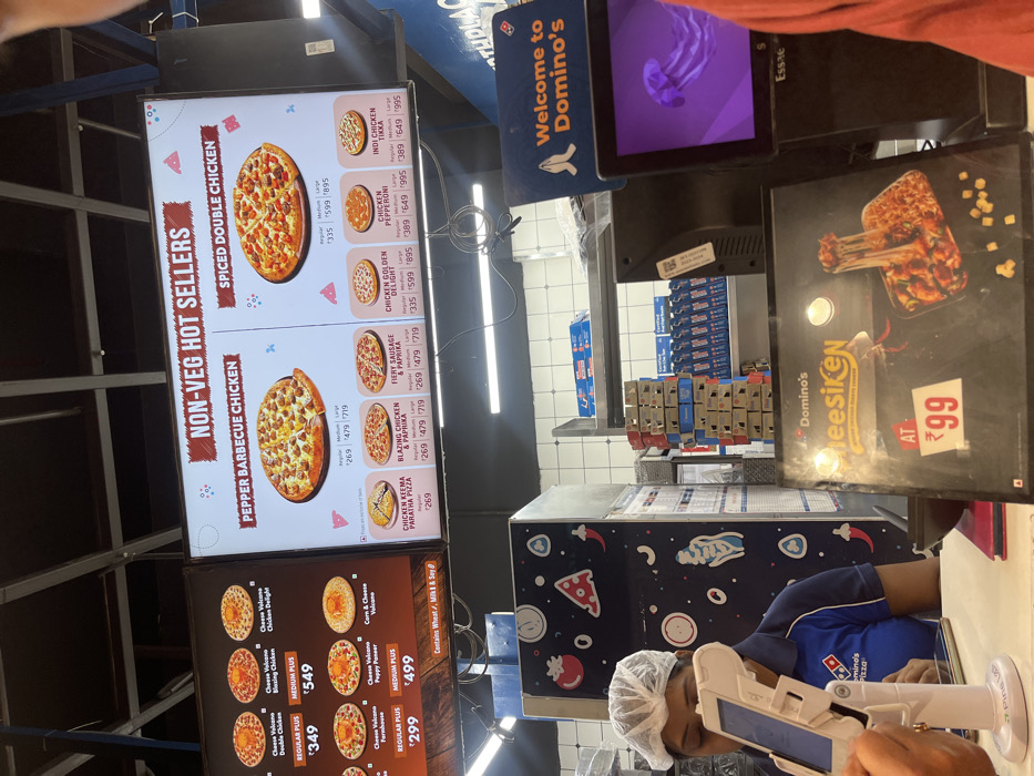 The image provides a snapshot inside a Domino's pizza outlet in Tinsukia, India, focusing on the service counter and menu displays. A Domino's employee, clad in a blue uniform and hairnet, is seen attending to a tablet device, suggesting they are processing an order or managing sales.
To the left, two brightly lit menu boards showcase a variety of pizzas. The first board, titled "NON-VEG HOT SELLERS," lists options such as "PEPPER BARBECUE CHICKEN" (₹269-₹719), "SPICED DOUBLE CHICKEN" (₹335-₹895), "CHICKEN KEEMA PARATHA PIZZA" (₹269), "BLAZING CHICKEN & PAPRIKA" (₹269-₹719), "FIERY SAUSAGE & PAPRIKA" (₹269-₹719), "CHICKEN GOLDEN DELIGHT" (₹335-₹895), "CHICKEN PEPPERONI" (₹389-₹995), and "INDI CHICKEN TIKKA" (₹389-₹995). The second menu board features "Cheese Volcano" pizzas, including "Double Chicken" (REGULAR PLUS ₹349), "Blazing Chicken" (MEDIUM PLUS ₹549), "Farmhouse" (REGULAR PLUS ₹299), and "Puppy Paneer" (MEDIUM PLUS ₹499), with an ingredient disclaimer stating "Contains Wheat, Milk & Soy."
A digital screen above the counter displays "Welcome to Domino's." Adjacent to the employee, a promotional sign highlights "Domino's Cheesiken AT ₹99." In the background, stacks of pizza boxes are visible on shelving, contributing to the typical quick-service restaurant environment. The indoor setting is well-lit with fluorescent lights, suggesting active business hours. The employee appears focused on their task, embodying the operational aspect of the restaurant.