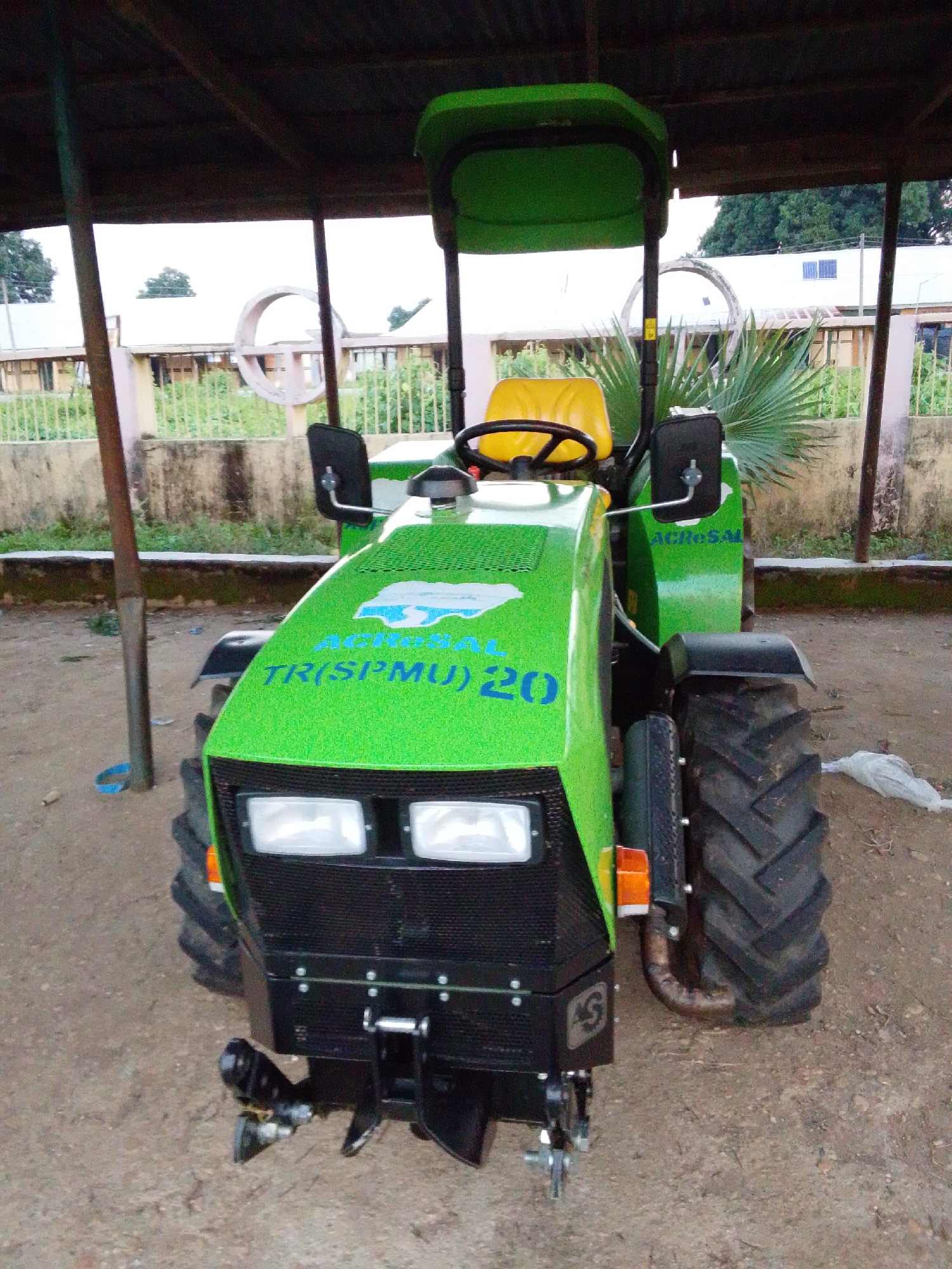 A bright green tractor is parked under a wooden shelter in Serti, Nigeria. The tractor has a yellow seat, a steering wheel, and large headlights. Text on the hood of the tractor reads "AGROSAL TR(SPMU) 20." The shelter has a corrugated metal roof and wooden supports. In the background, a building with a white roof and a fence can be seen. The ground is covered in dirt and gravel.