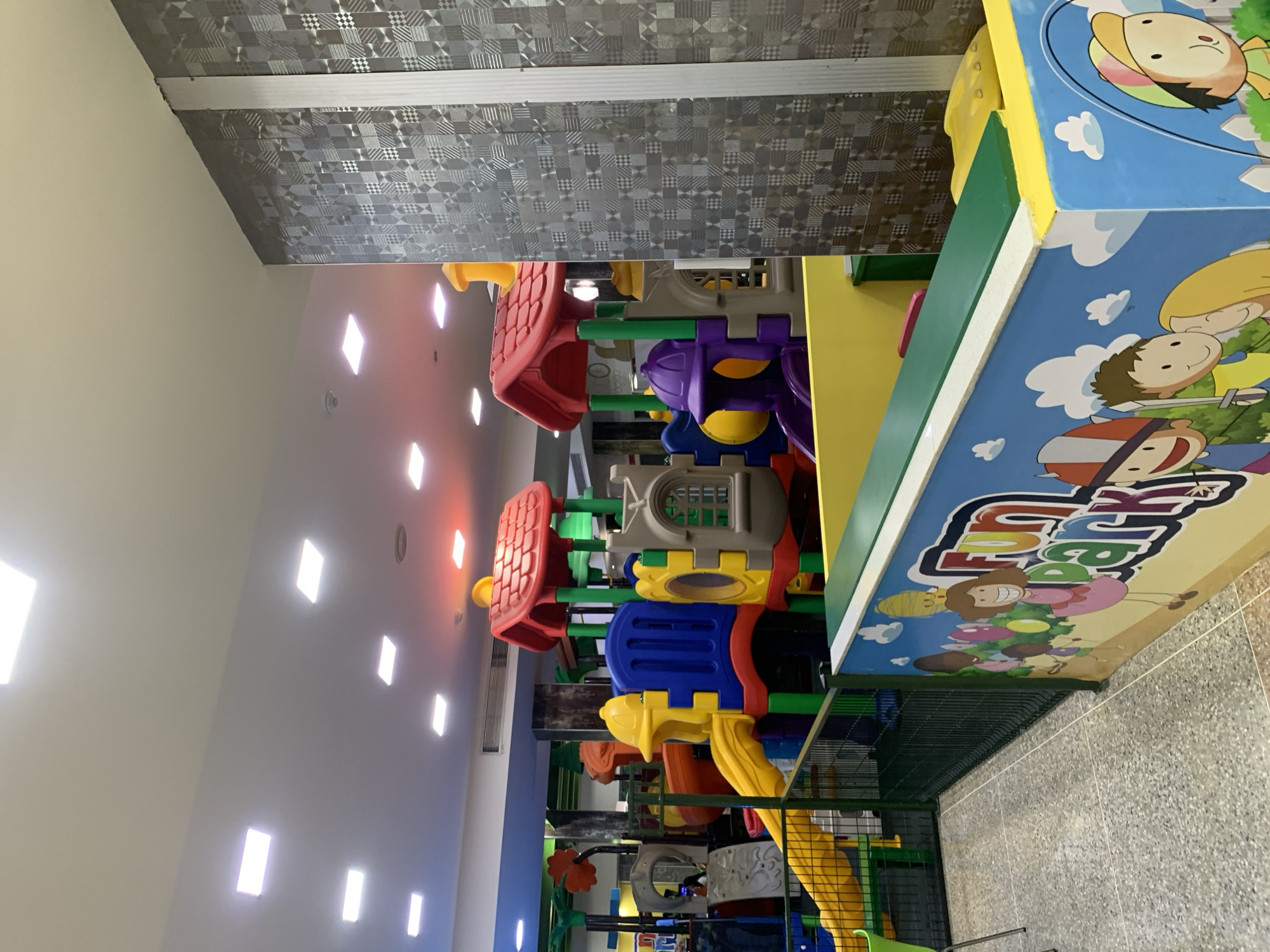 The image depicts an indoor children's play area, likely within a commercial establishment in El Valle del Espíritu Santo, Venezuela. The foreground features a brightly colored, low-rise play structure with slides, tunnels, and climbing elements made of plastic and metal. The structure is predominantly red, yellow, blue, and purple. A green safety fence encloses part of the play area. The lower section of the enclosure is decorated with cartoon characters and the words "Fun Park" in vibrant colors. The floor is tiled with a speckled pattern. In the background, more play structures are visible, along with overhead lighting panels in the ceiling. A patterned wall is present on the right side of the image. No people are clearly visible in the frame.
