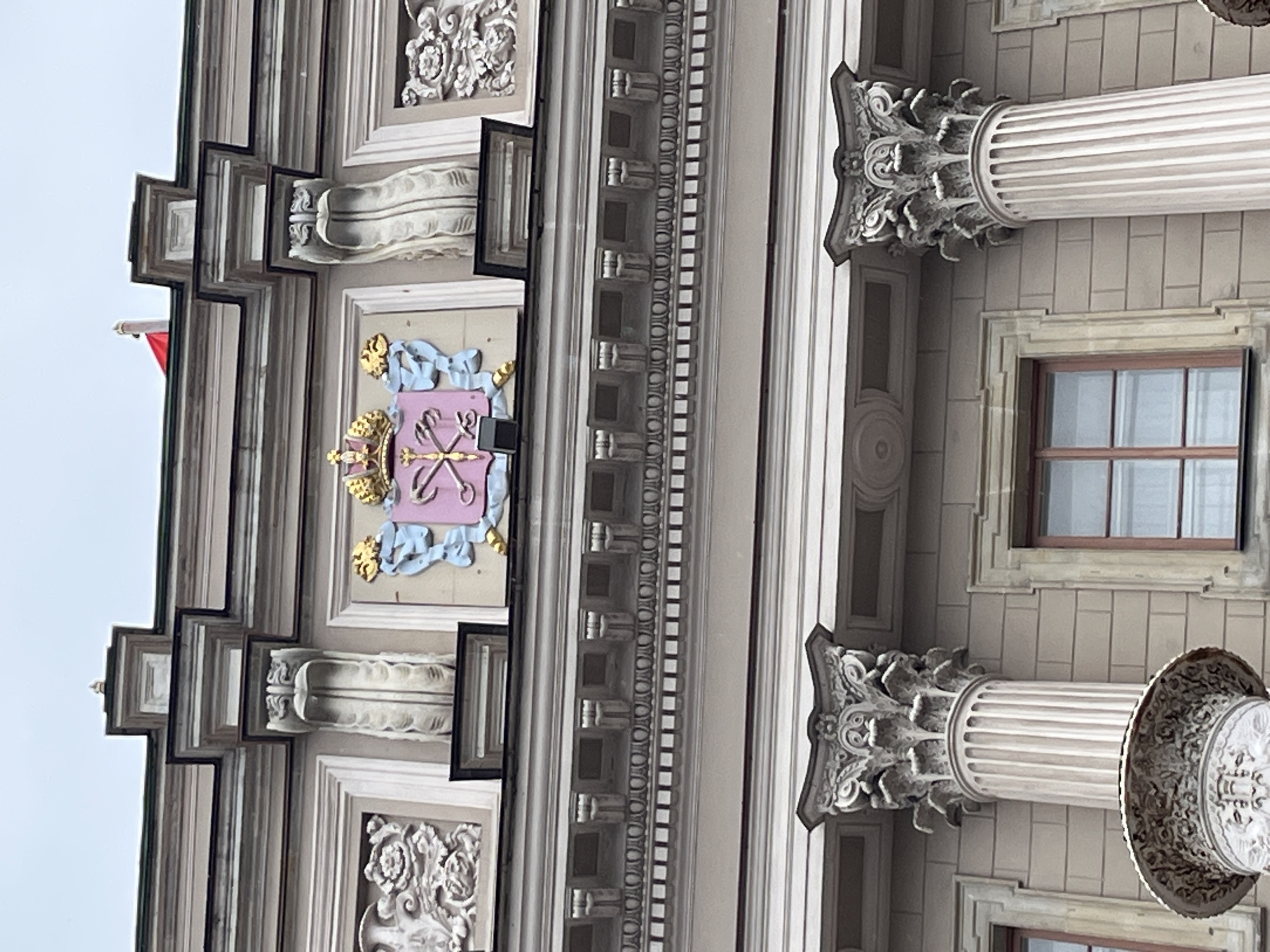 The image captures a portion of a building facade in Saint Petersburg, Russia, likely a historical or administrative building due to its ornate architectural details. The building is predominantly gray. It features classical elements such as columns with Corinthian capitals, decorative moldings, framed windows, and a coat of arms. The coat of arms, located centrally, is pink with gold and blue embellishments. A small red flag is visible on the left side of the facade. The sky is overcast.