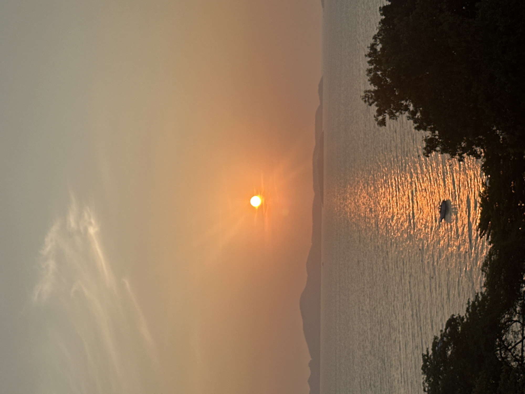 The image depicts a sunset over the ocean in Pattaya City, Thailand. The sun is positioned near the center of the frame, casting a bright, orange glow across the sky and reflecting off the water. The coastline is visible in the distance as a dark line. Trees are located to the right, and a single small boat can be seen on the water.